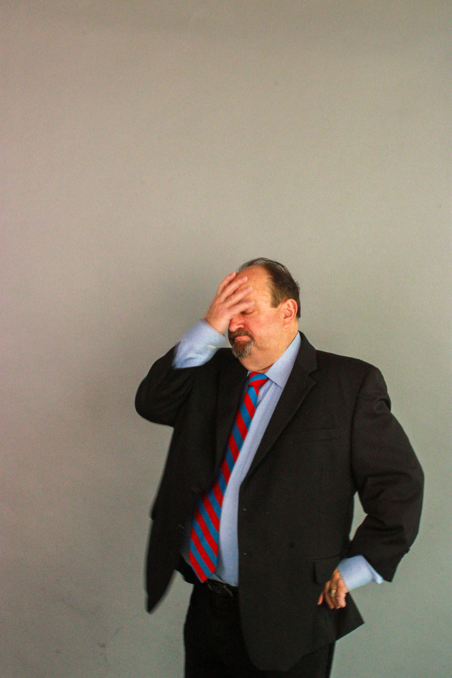 A person in a business suit and red tie gestures with hand covering face in a dramatic pose.