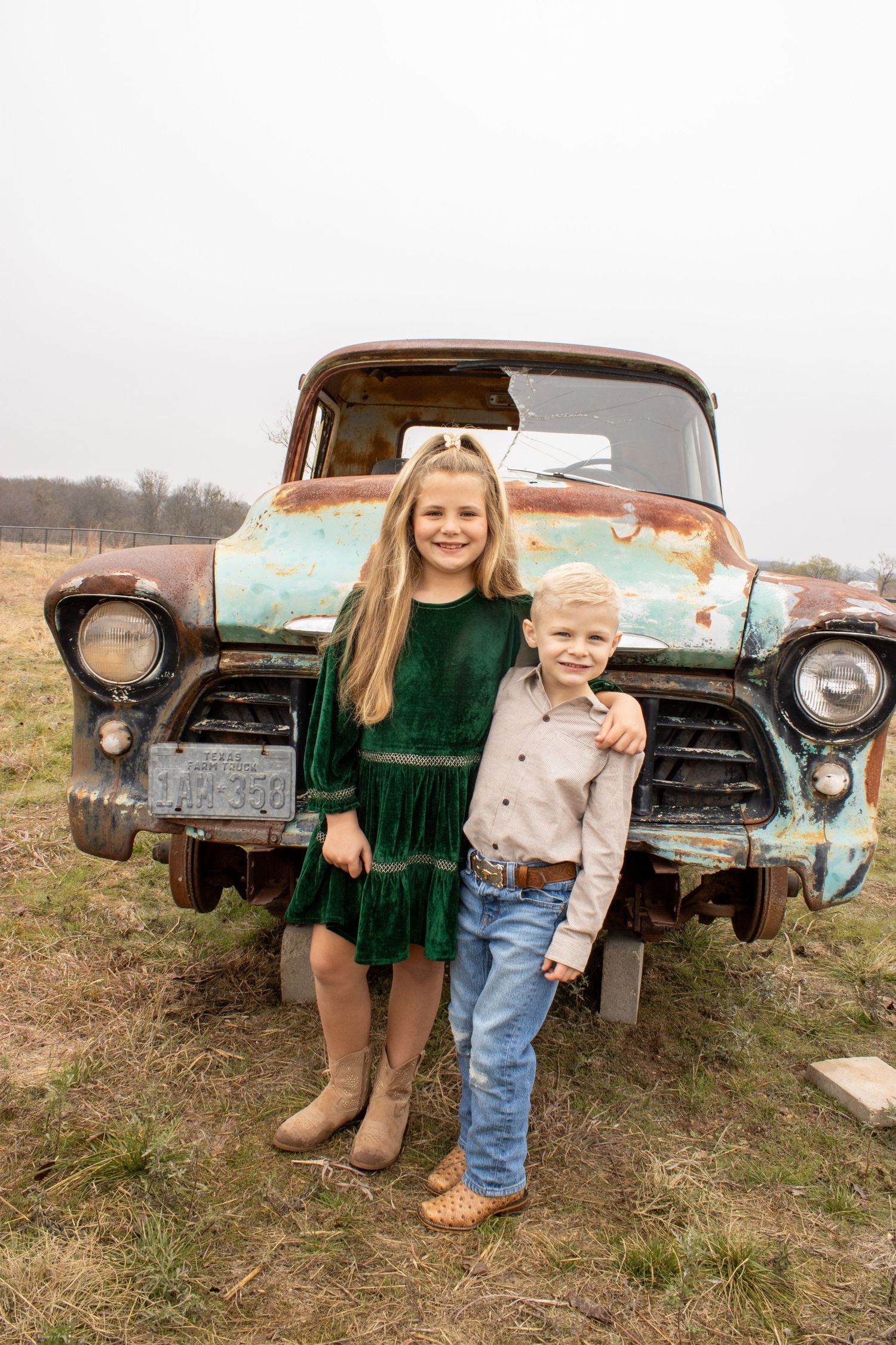 Two children pose in front of a rustic vintage truck in a grassy field wearing contrasting green and beige outfits.