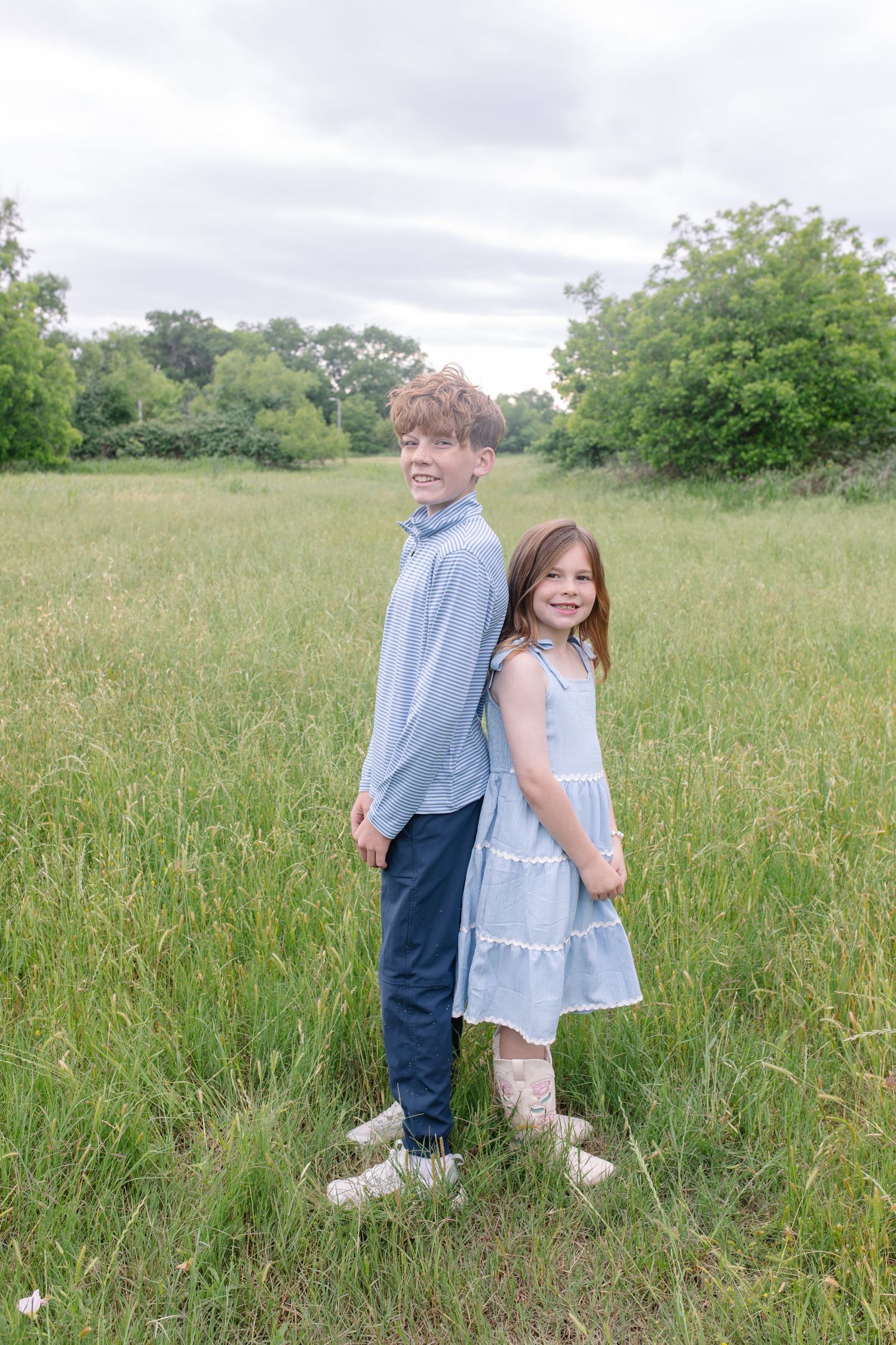 Two children in blue outfits stand back-to-back smiling in a lush green meadow on a cloudy day.
