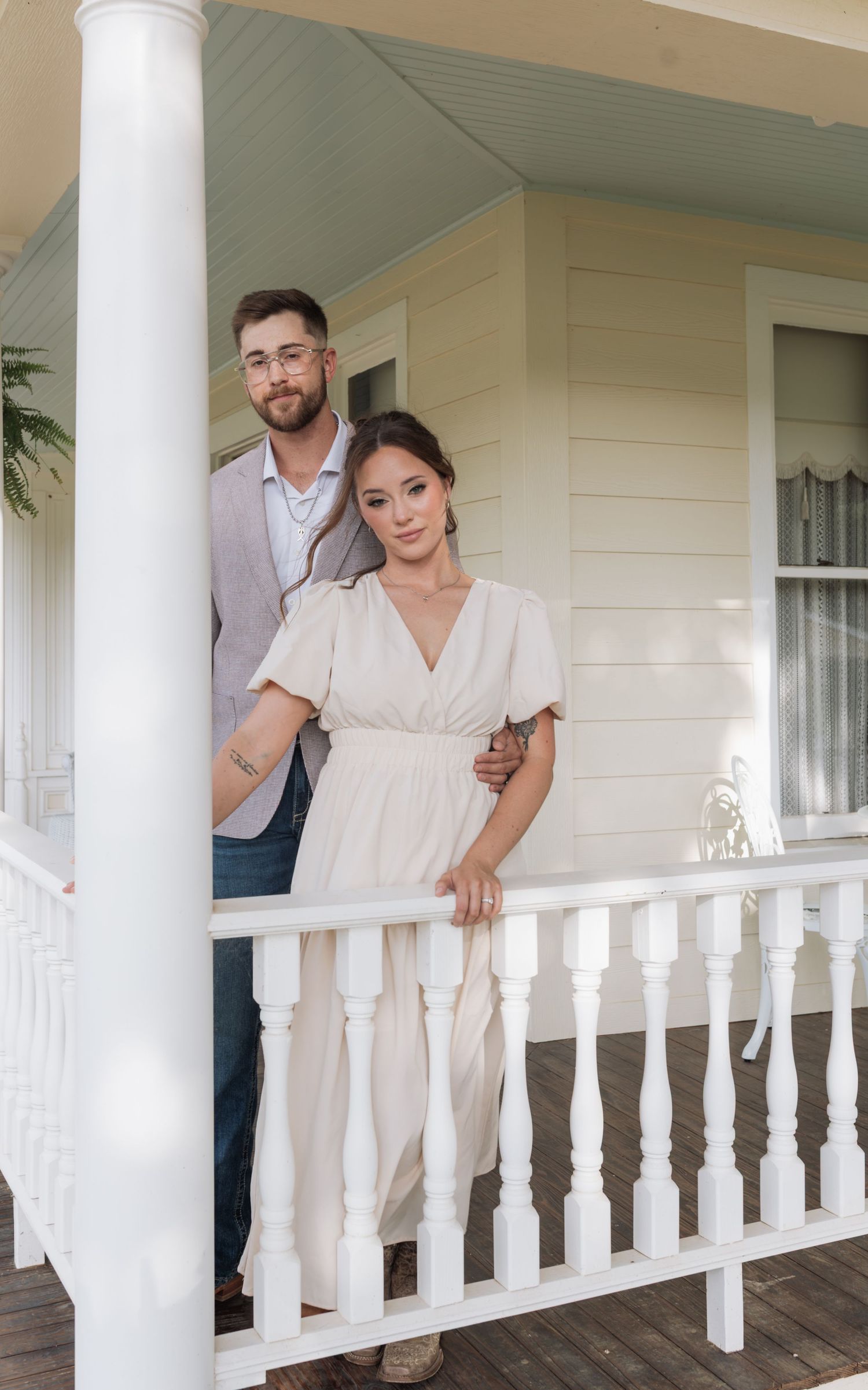 Couple posing on a white railed porch, woman in cream dress, man in gray blazer and jeans, beside a yellow house.