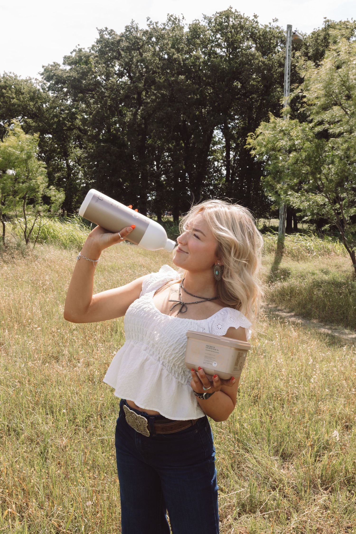 Blonde woman in white top drinks from tumbler while holding box in sunny green field.