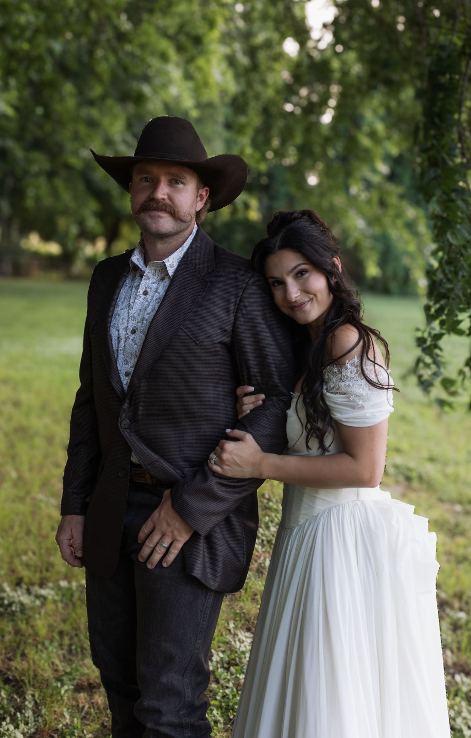 Couple in western wedding attire poses outdoors, man in cowboy hat and suit, woman in white bridal dress, green trees background.