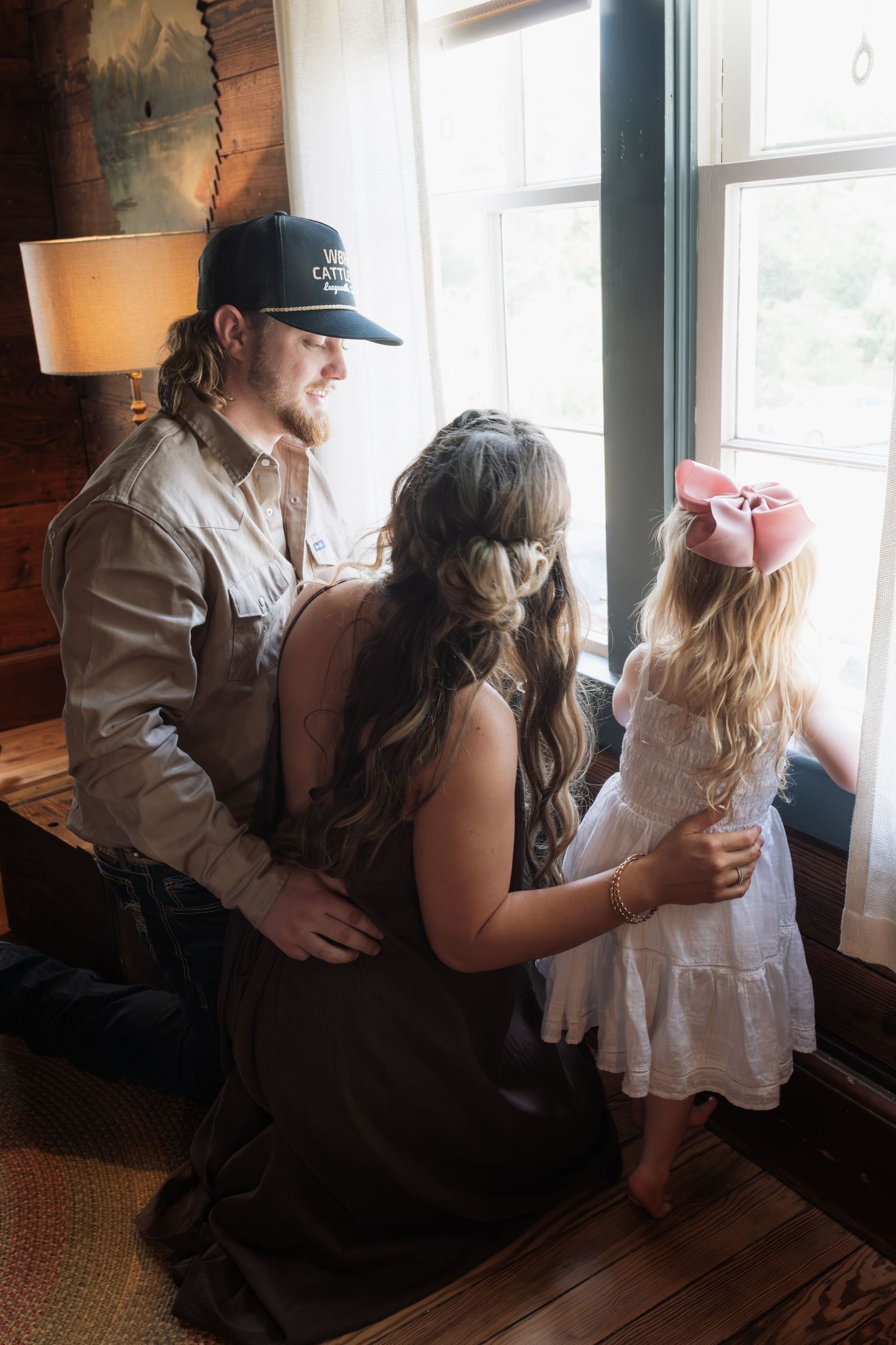 A man and woman kneel beside a young girl in a pink bow looking out a rustic cabin window together.