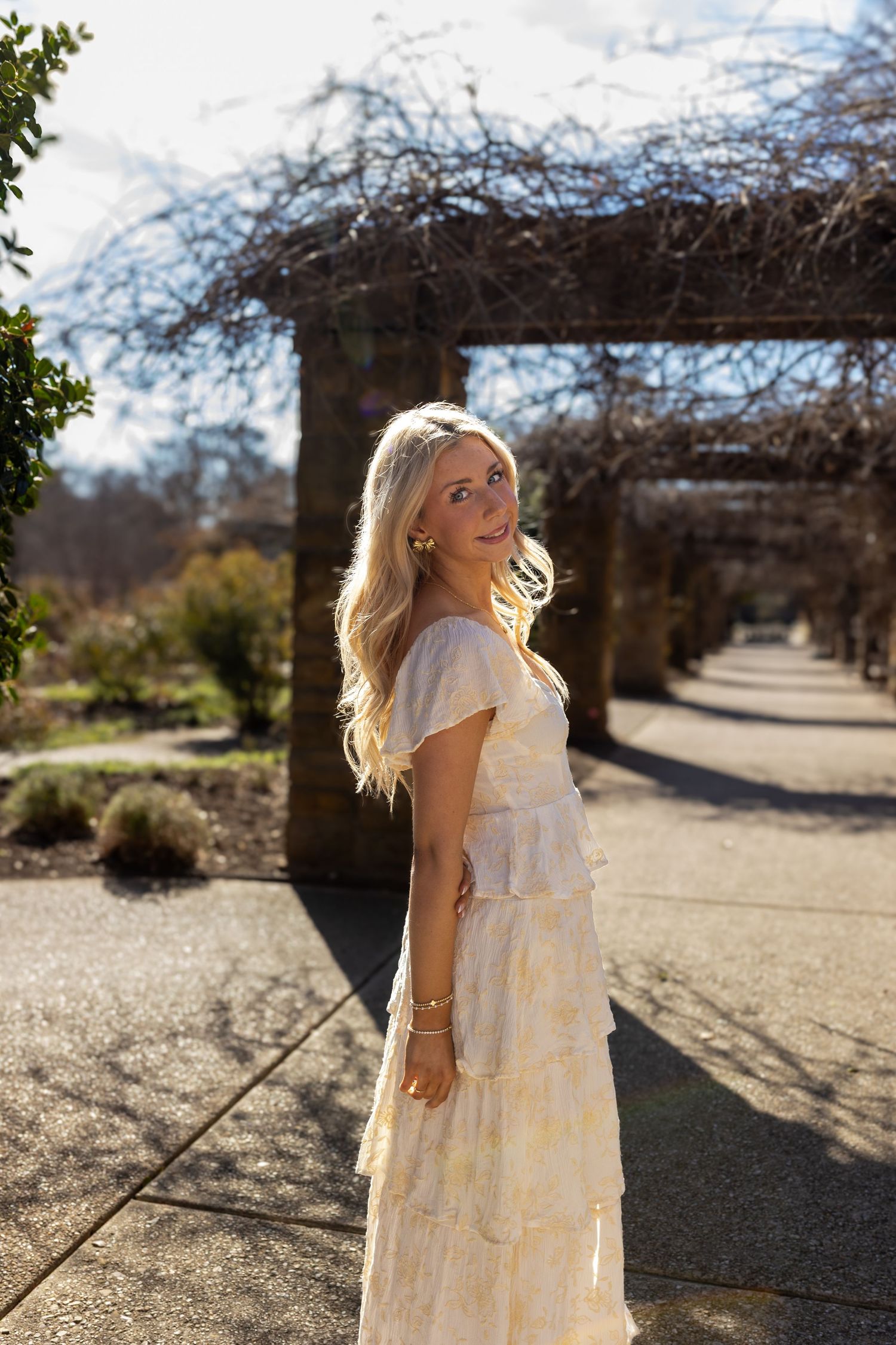 A woman in a flowing white tiered dress stands in a sunlit garden path with bare trees in the background.