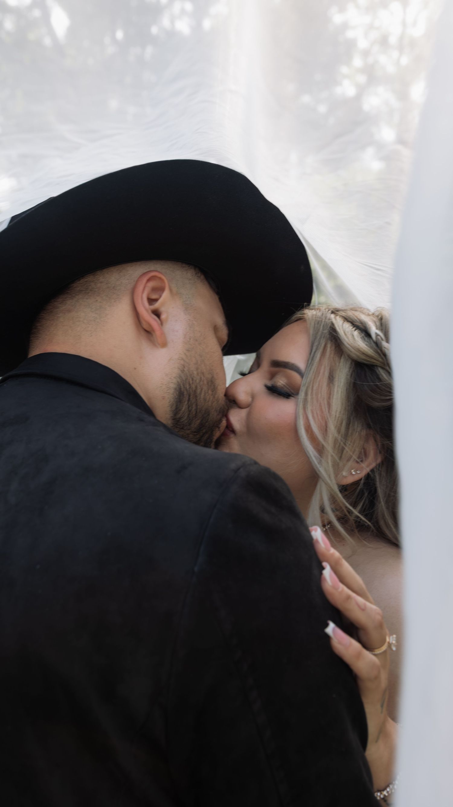 A bride and groom sharing a romantic kiss, with the groom wearing a black cowboy hat at their wedding.