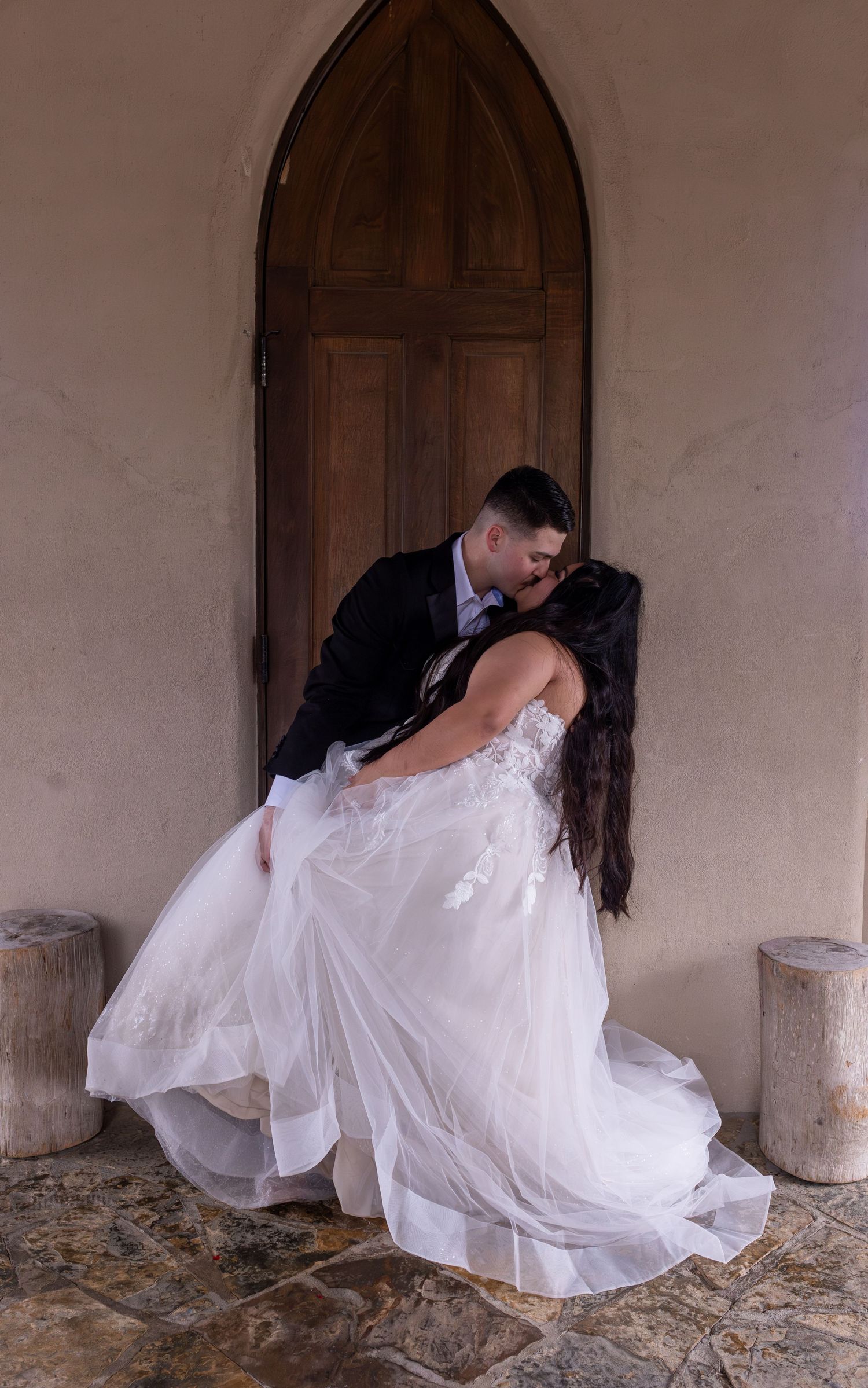 A romantic wedding couple shares a tender dip kiss pose in a rustic stone archway setting.