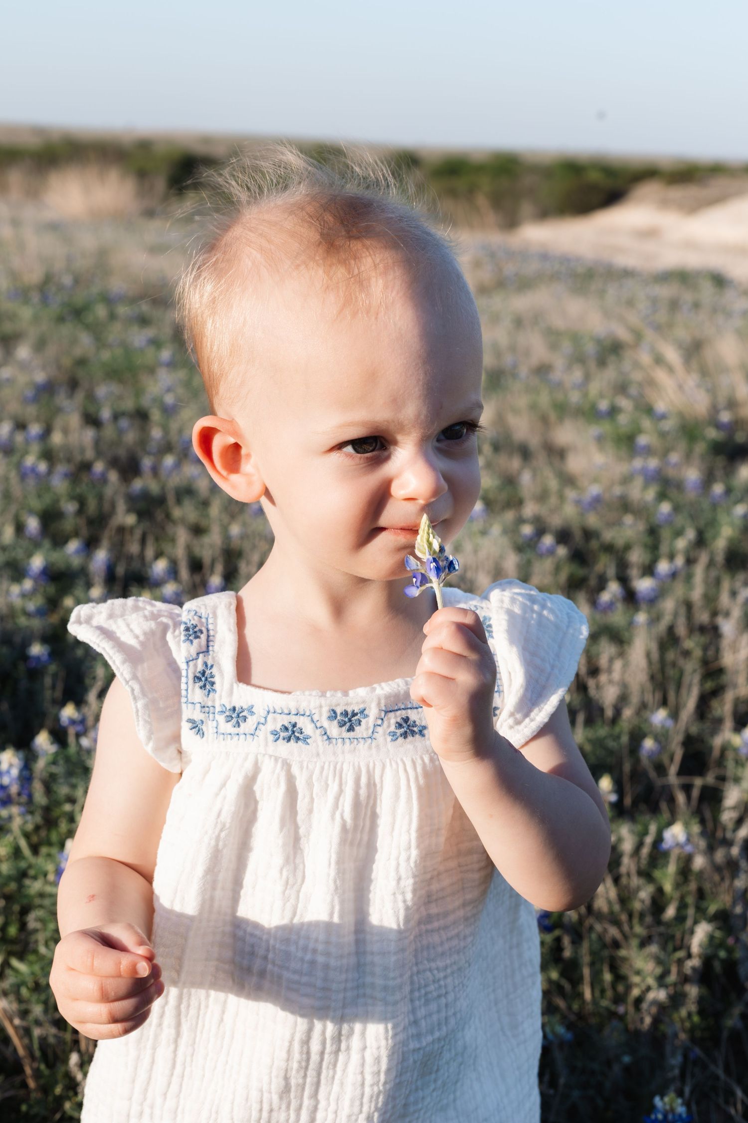 A young child in a white lace dress stands in a field of wildflowers during golden hour.