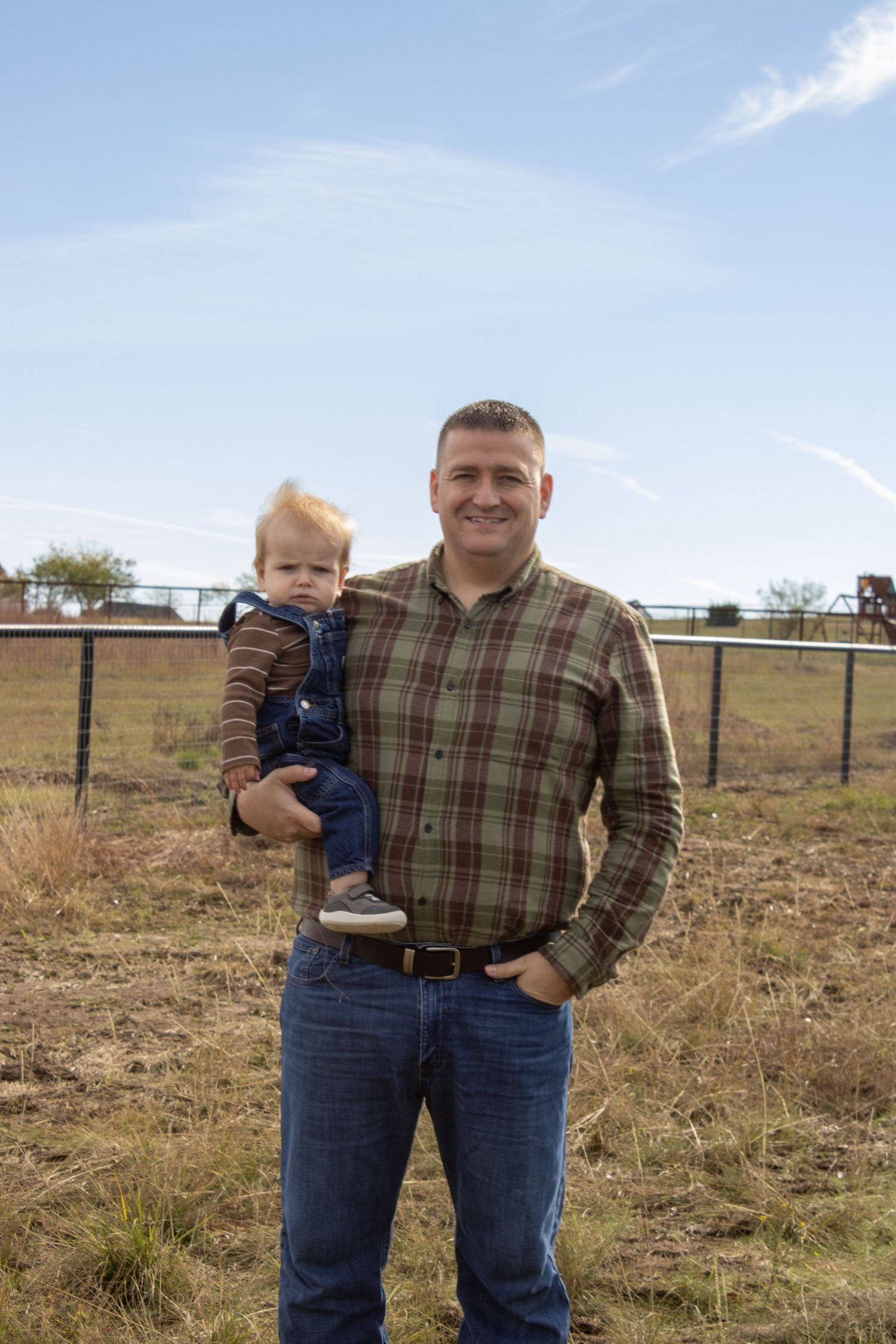 Adult and small child standing together in a grassy field with wooden fencing visible in the background.