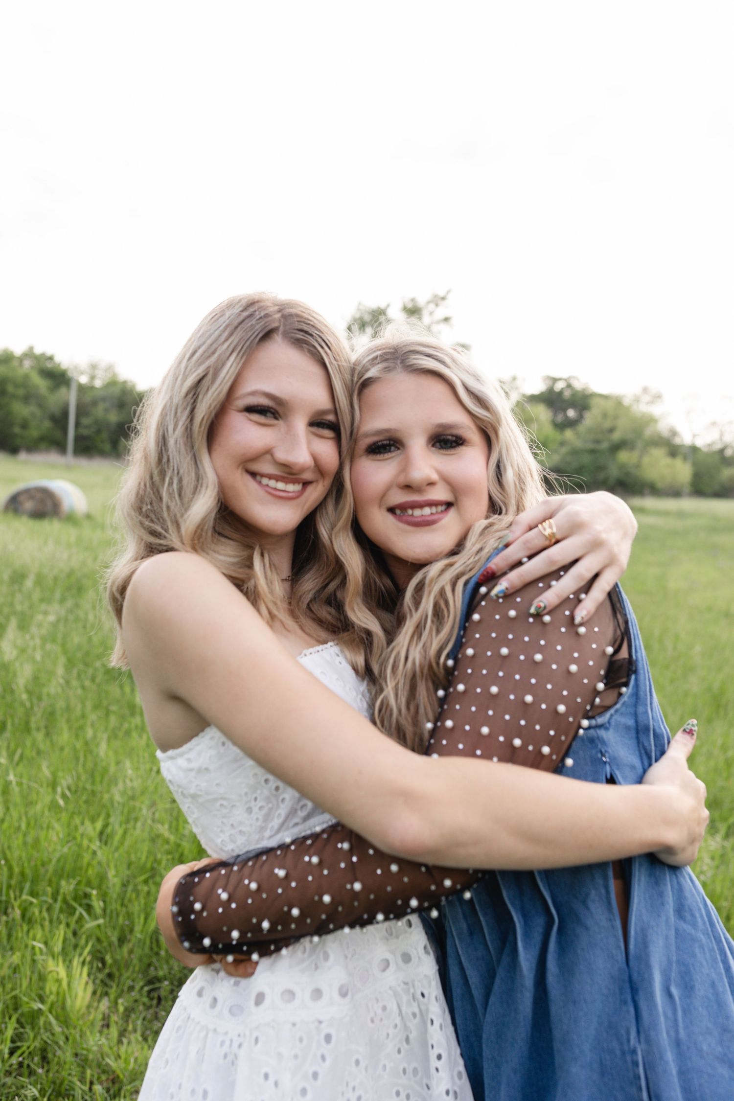 Two blonde young women embracing and smiling outdoors in a lush green field.
