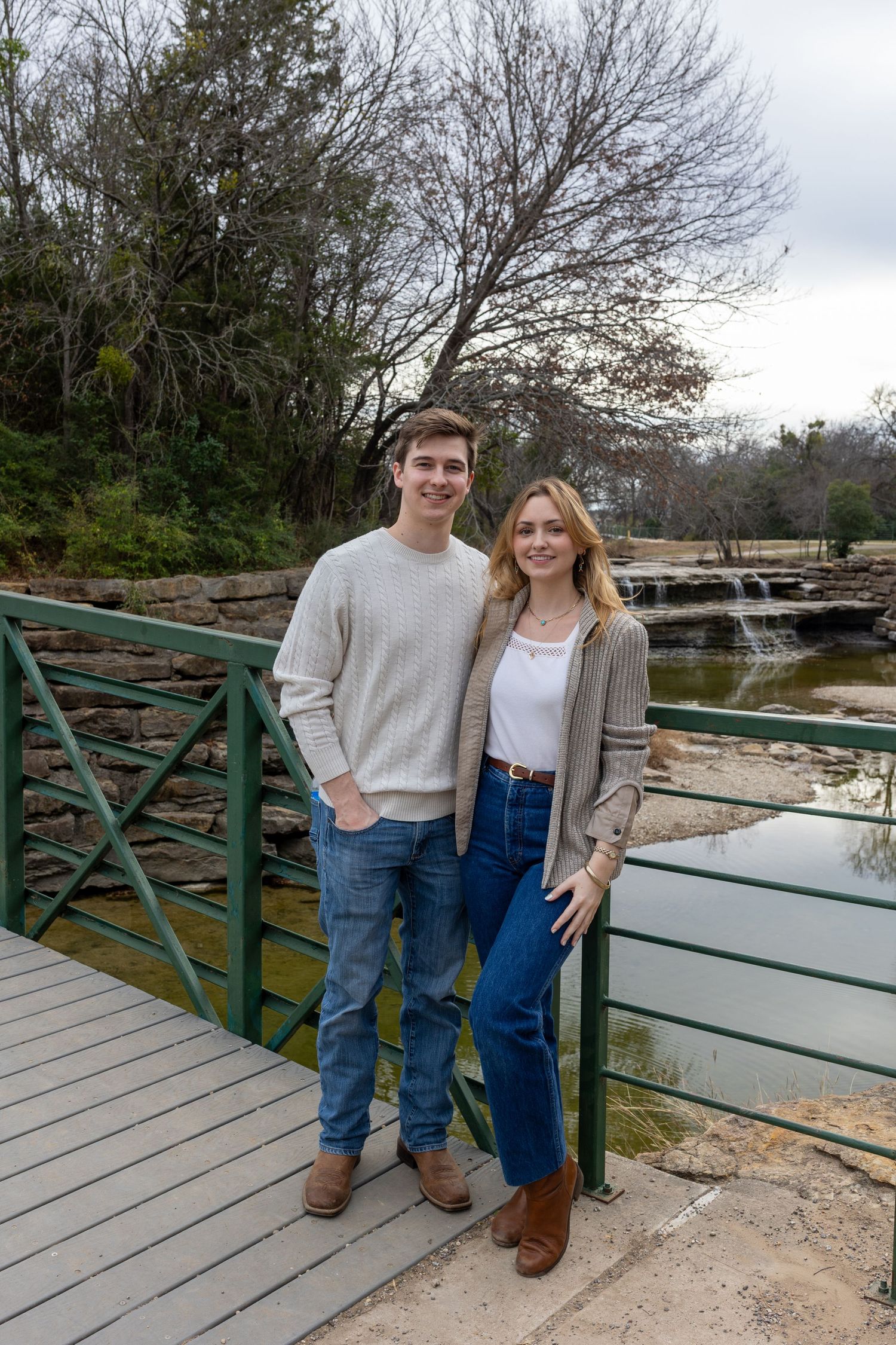 A couple stands together on a wooden bridge overlooking a scenic waterway on a cloudy winter day.