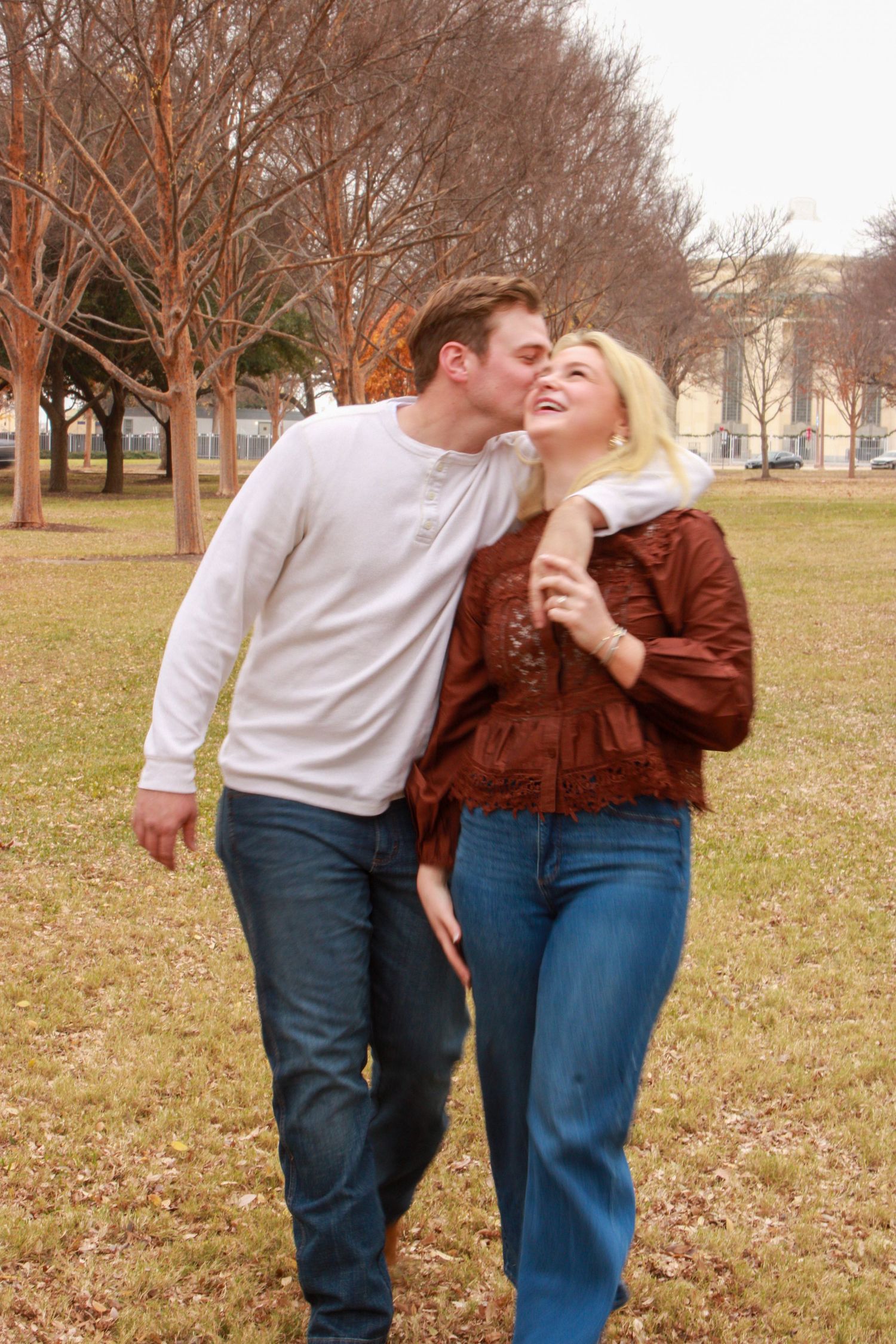 A couple walks in a park, man kissing woman on cheek, both wearing jeans, surrounded by bare winter trees.