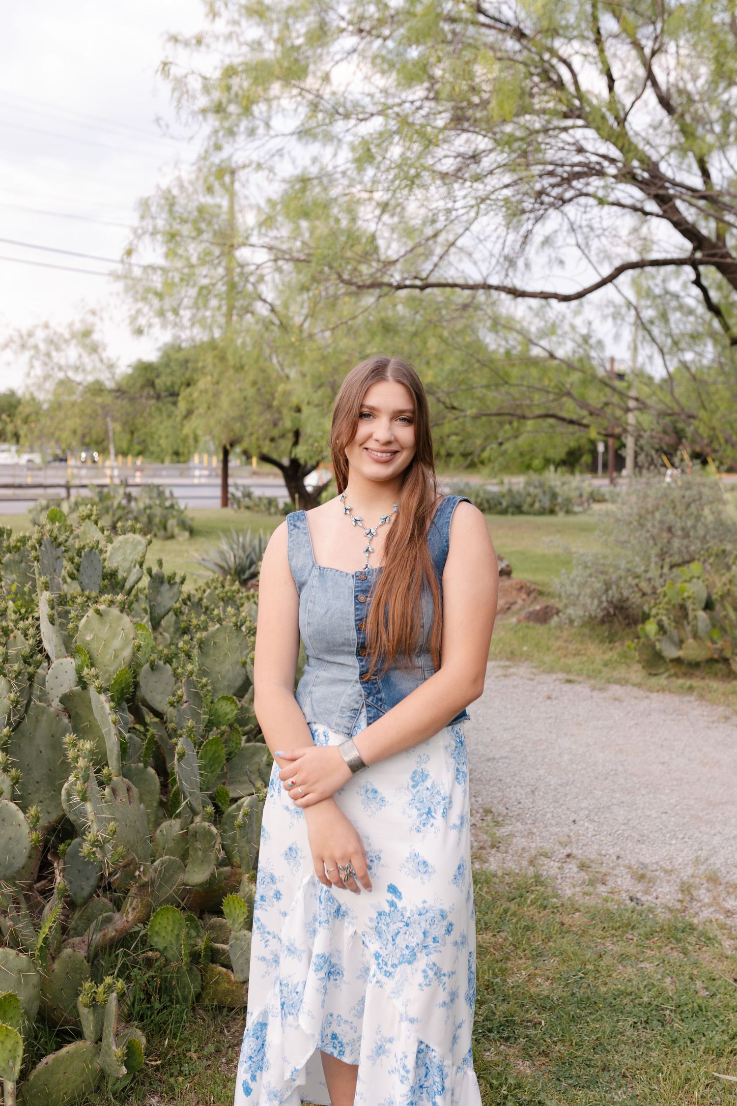 A person in a blue and white floral dress poses on a path with cactus plants and trees in the background.