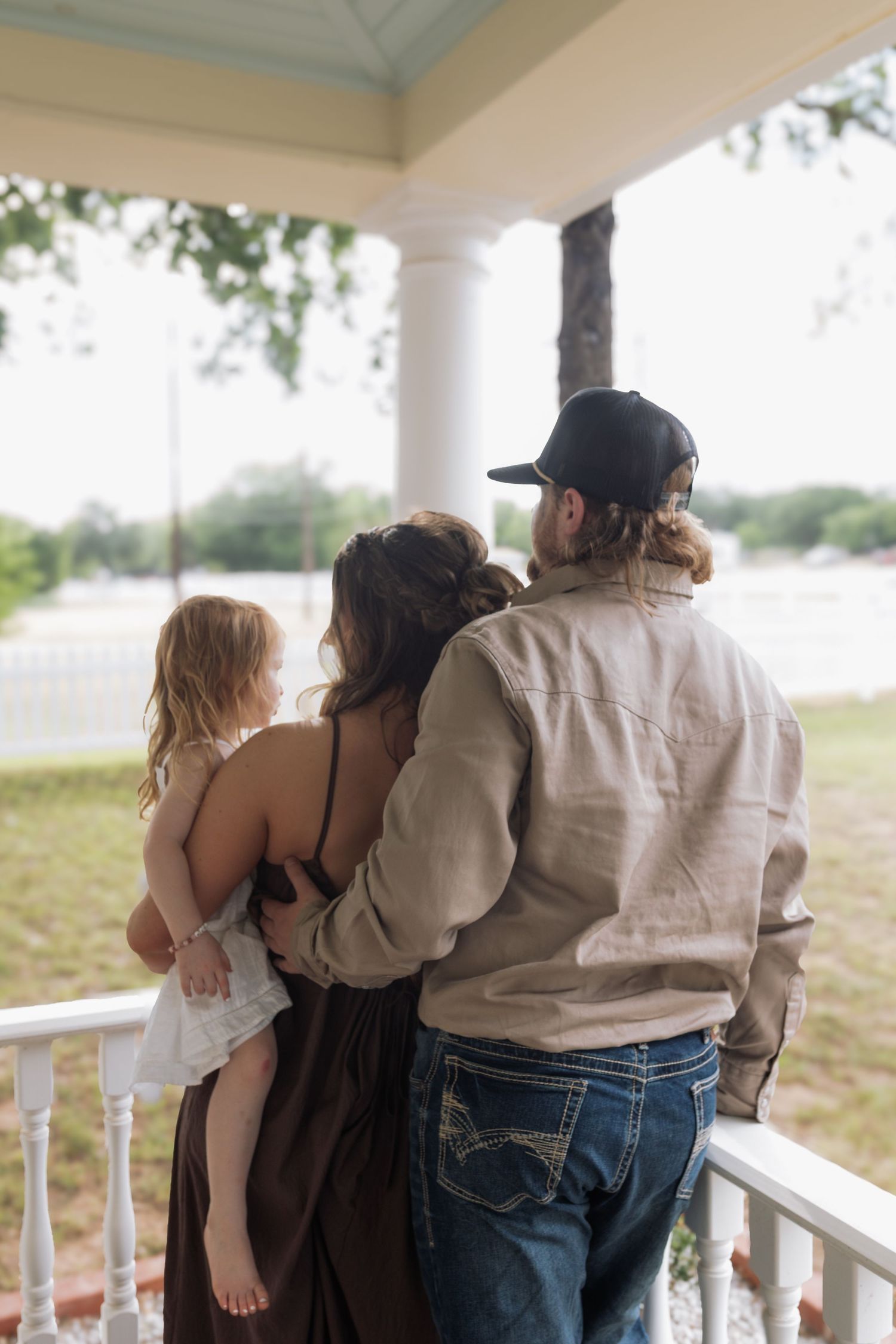 Family of three standing on a porch, parents holding young child, viewed from behind on a sunny day.