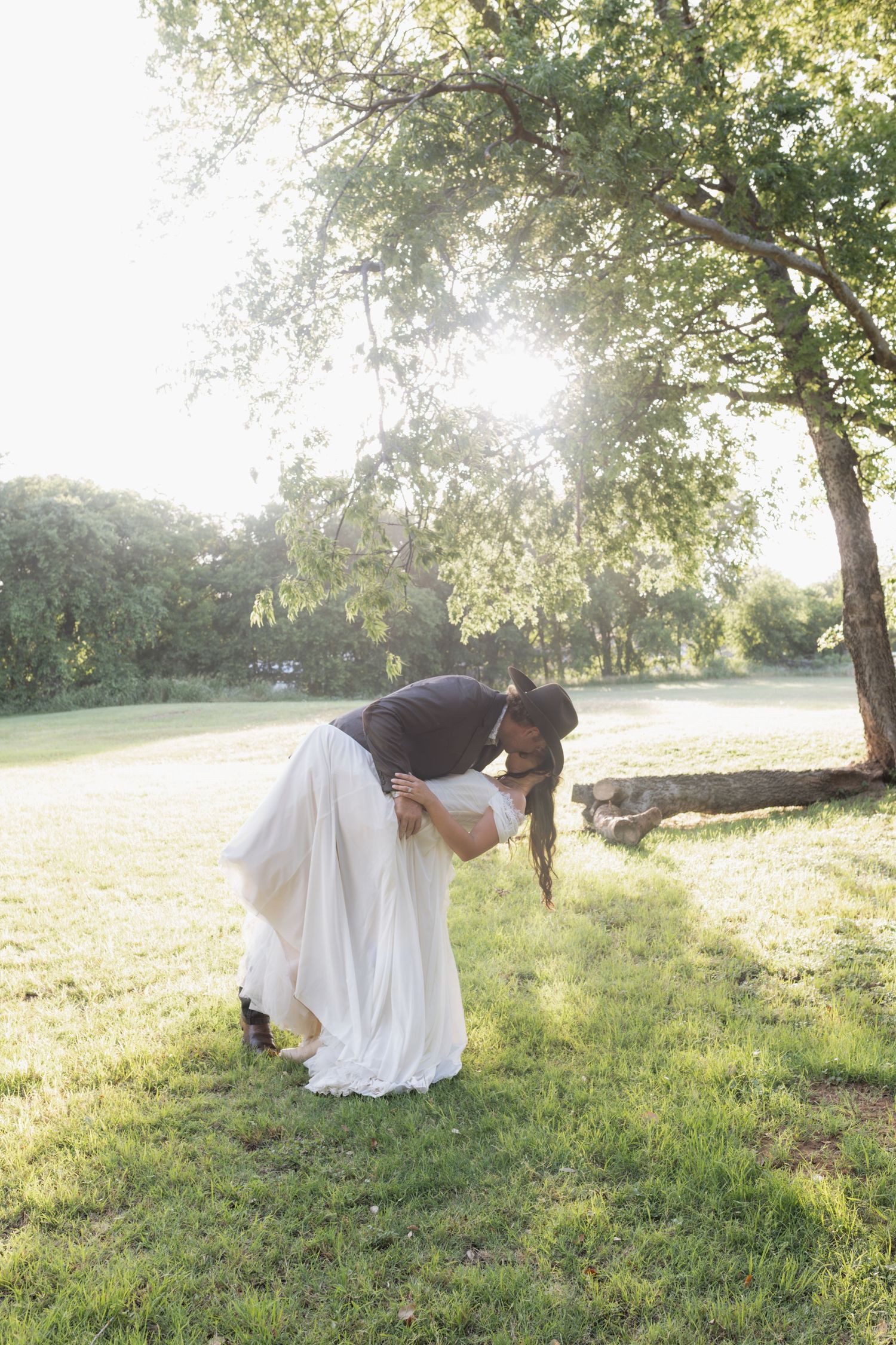 Couple sharing a romantic dip kiss on green grass under sunlit trees in outdoor wedding attire.