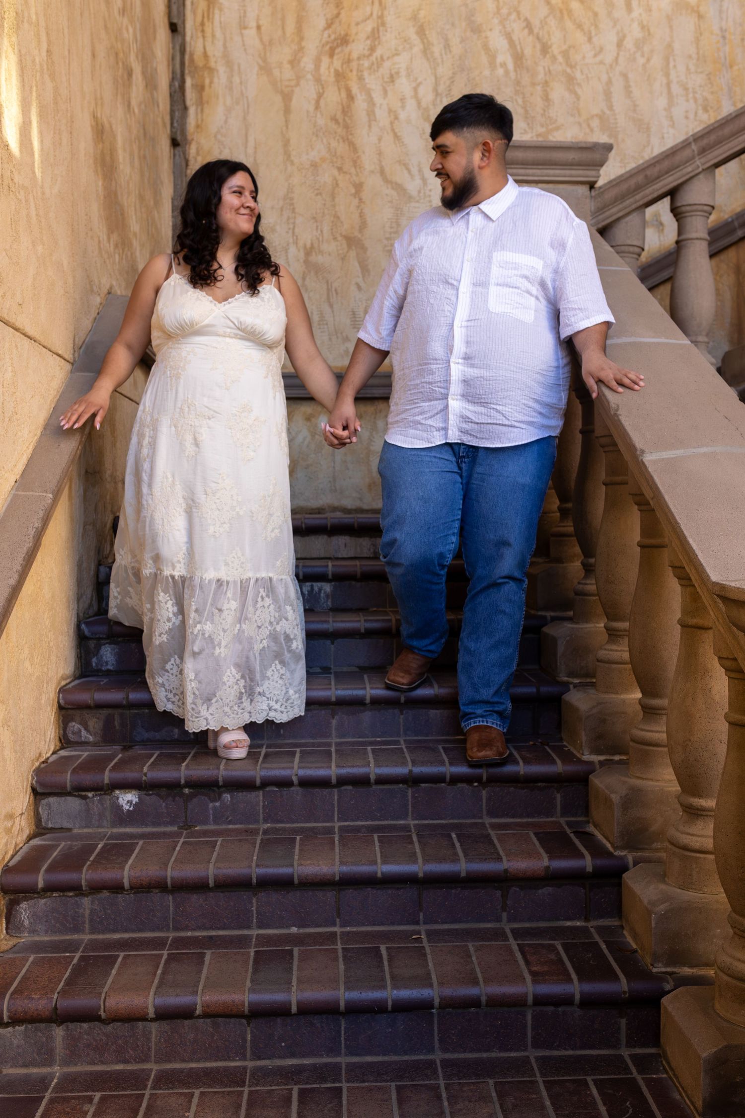 Couple in white and blue outfits holding hands while walking up stone stairs in a romantic engagement photo sequence.