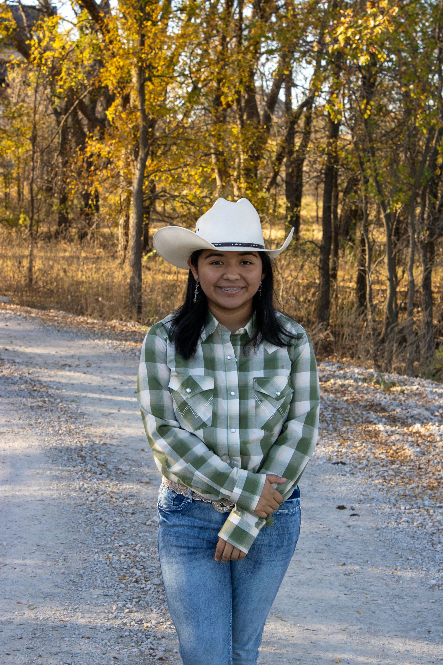 Someone in a green plaid shirt and cowboy hat poses on a dirt path surrounded by autumn trees with golden leaves.