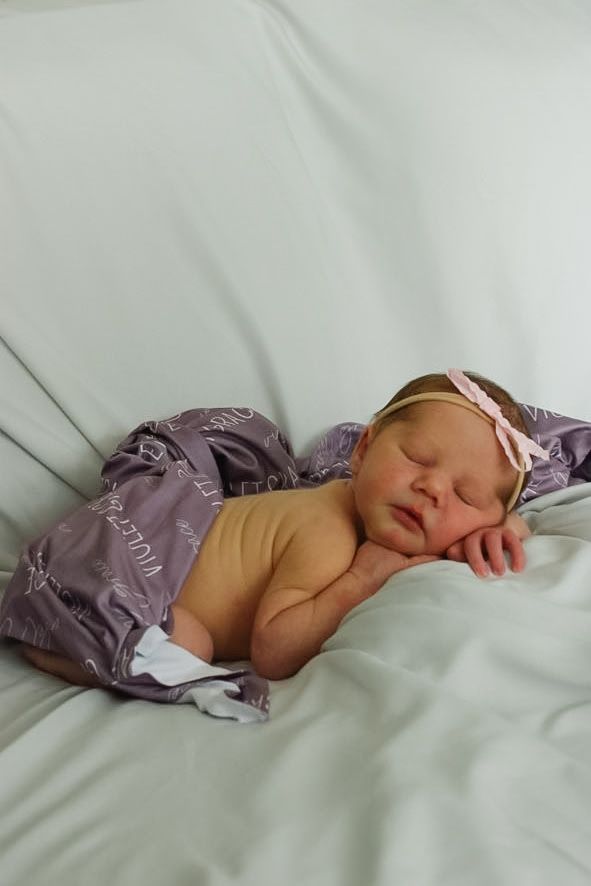 Sleeping newborn wrapped in purple blanket lying peacefully on white bedding.