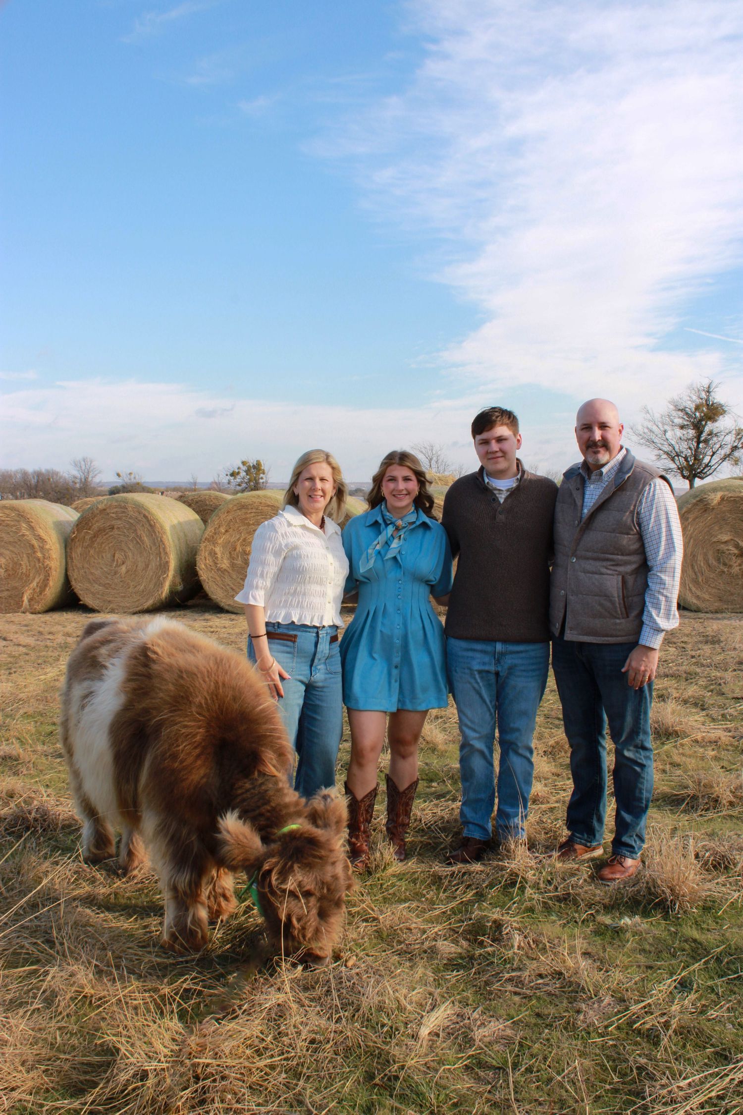 Group of people standing together in a field with hay bales and a brown pony grazing in the foreground.