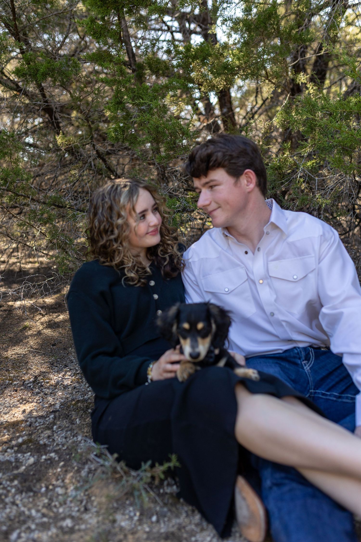 A couple sits together outdoors with their small black dog against a natural backdrop of trees and foliage.