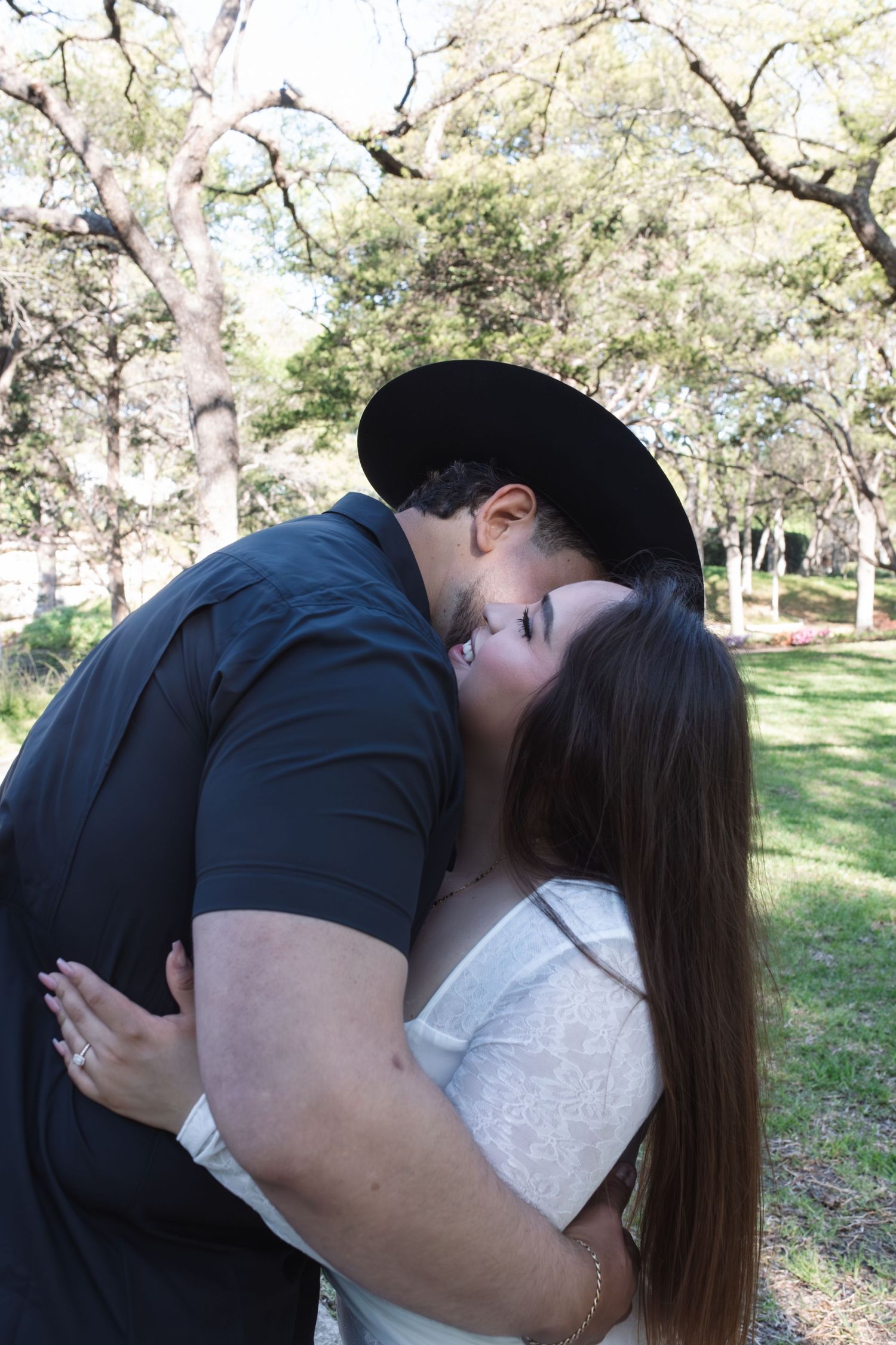 A couple embraces in a sunlit park setting with blooming trees in the background.