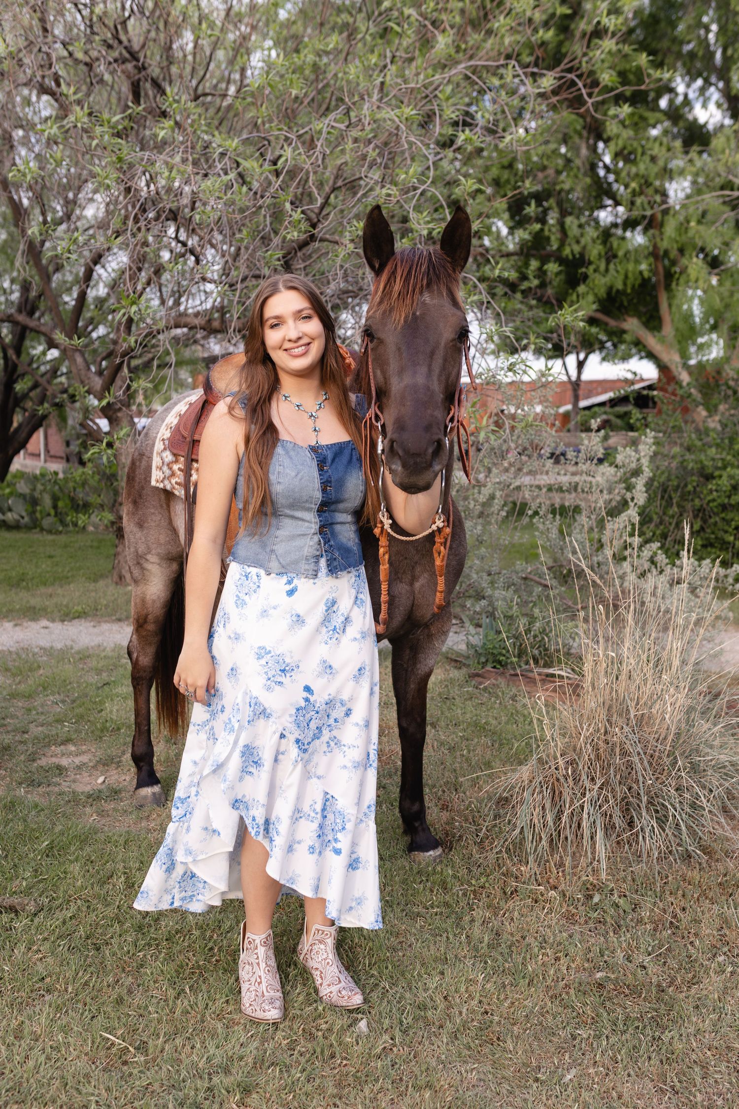 A woman in a floral dress poses with a dark brown horse in an outdoor setting with trees and grass.