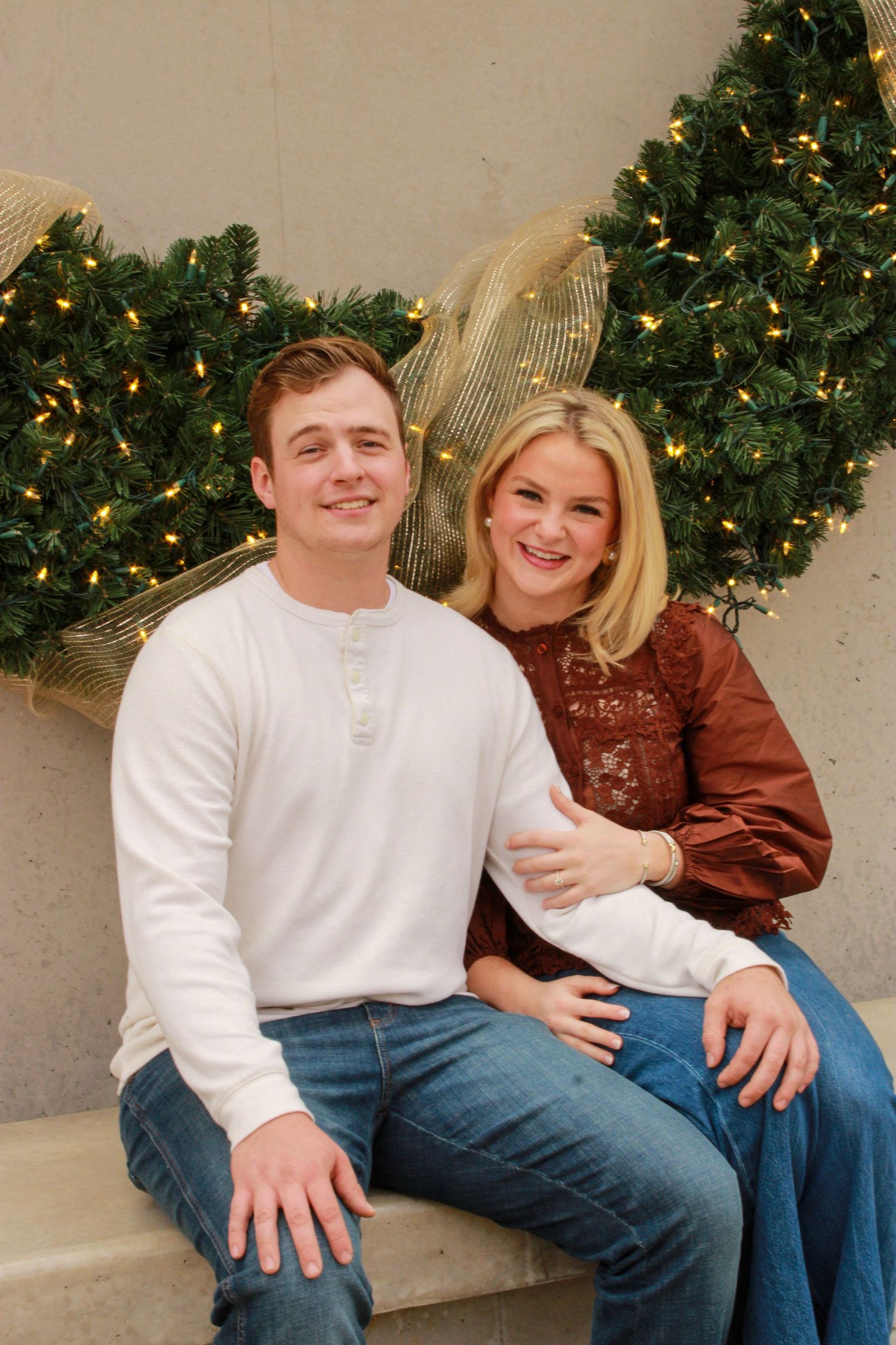 A smiling couple poses together in front of a decorated Christmas wreath with warm lights for a festive holiday portrait.
