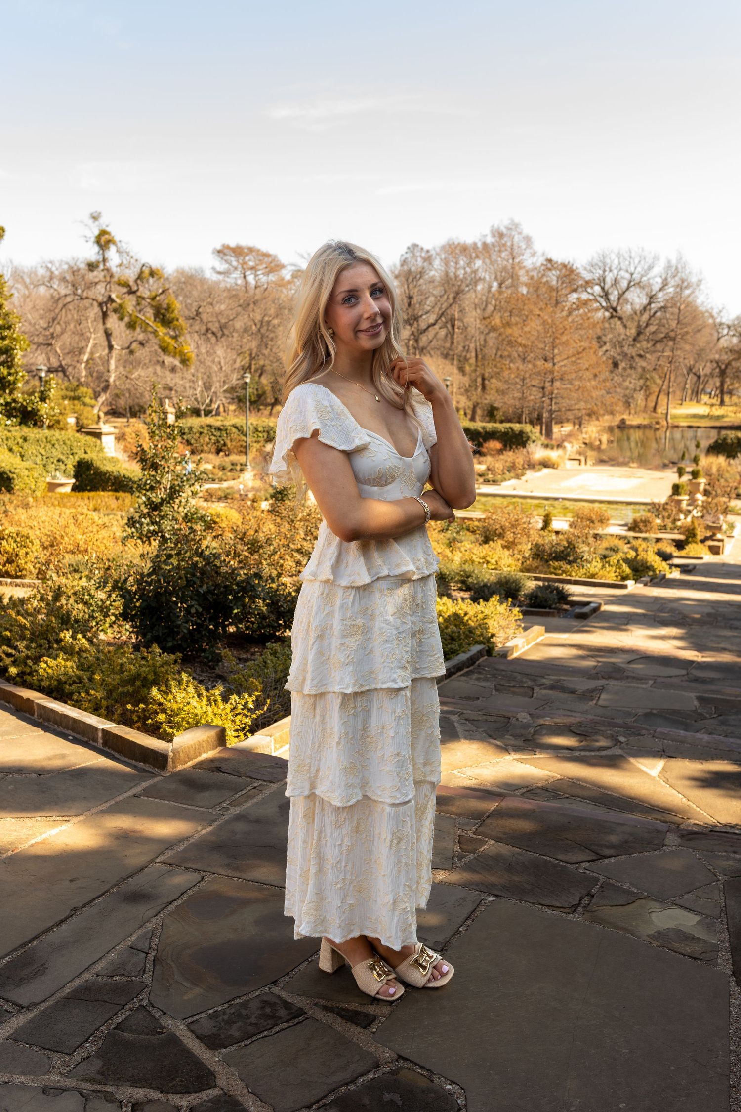 A person in a flowing white tiered dress poses in a garden setting with stone pathways during golden hour.