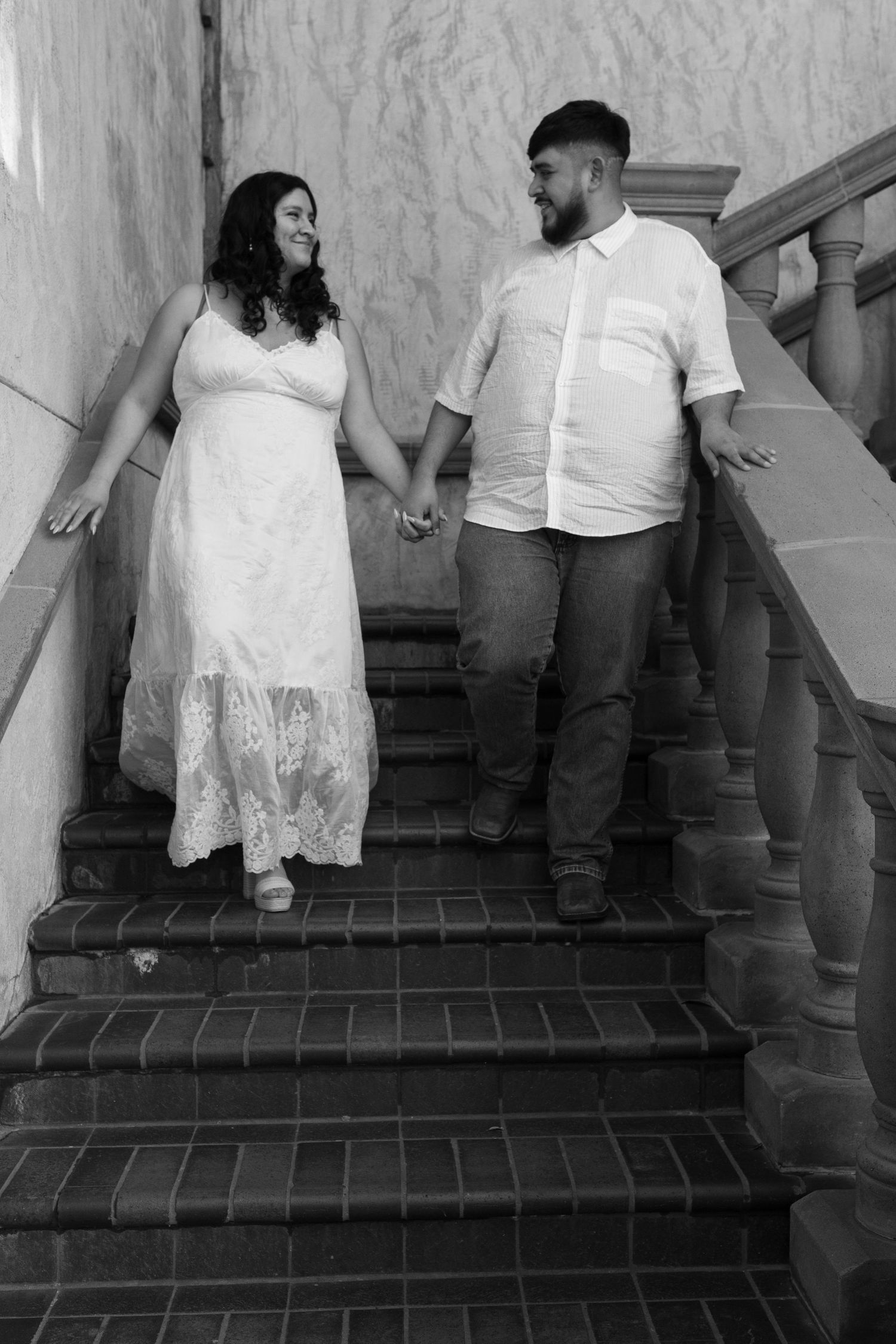 A couple in white attire hold hands while standing on brick stairs in a black and white photograph.