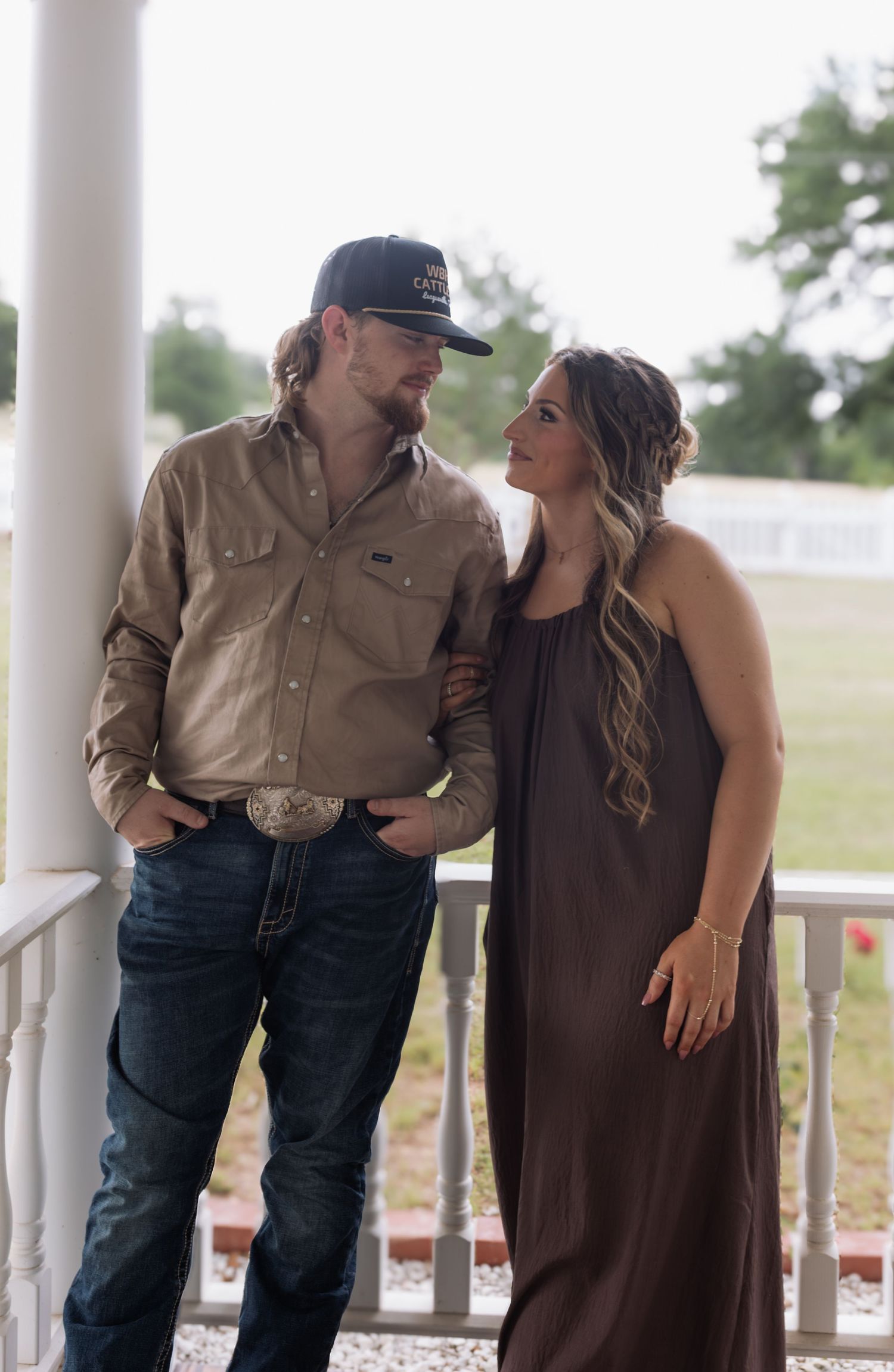 A couple in country attire poses on a white porch, man in tan shirt and jeans, woman in brown maxi dress.
