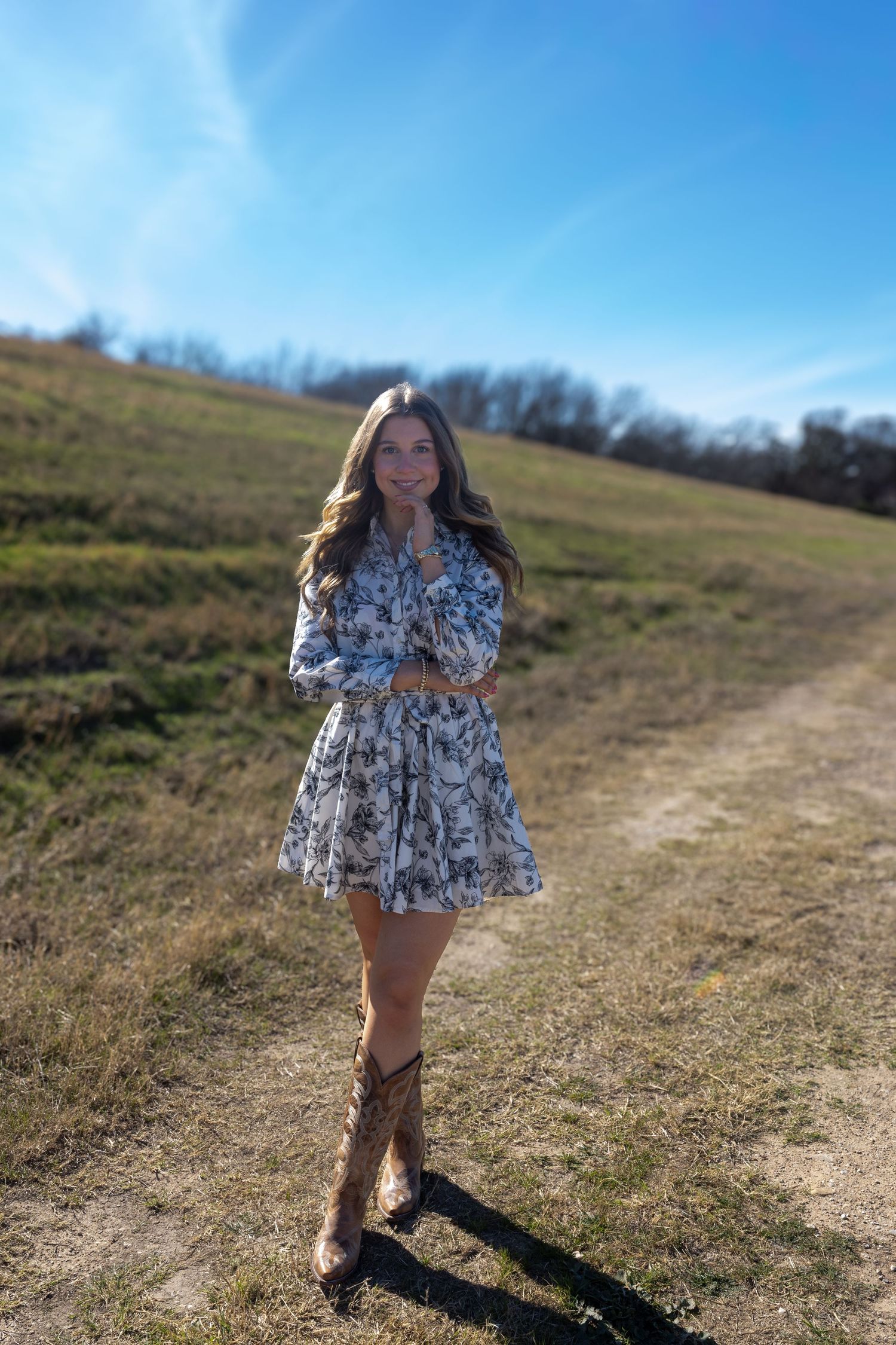 A figure in a floral print dress stands on a grassy hillside path against a bright blue sky on a sunny day.
