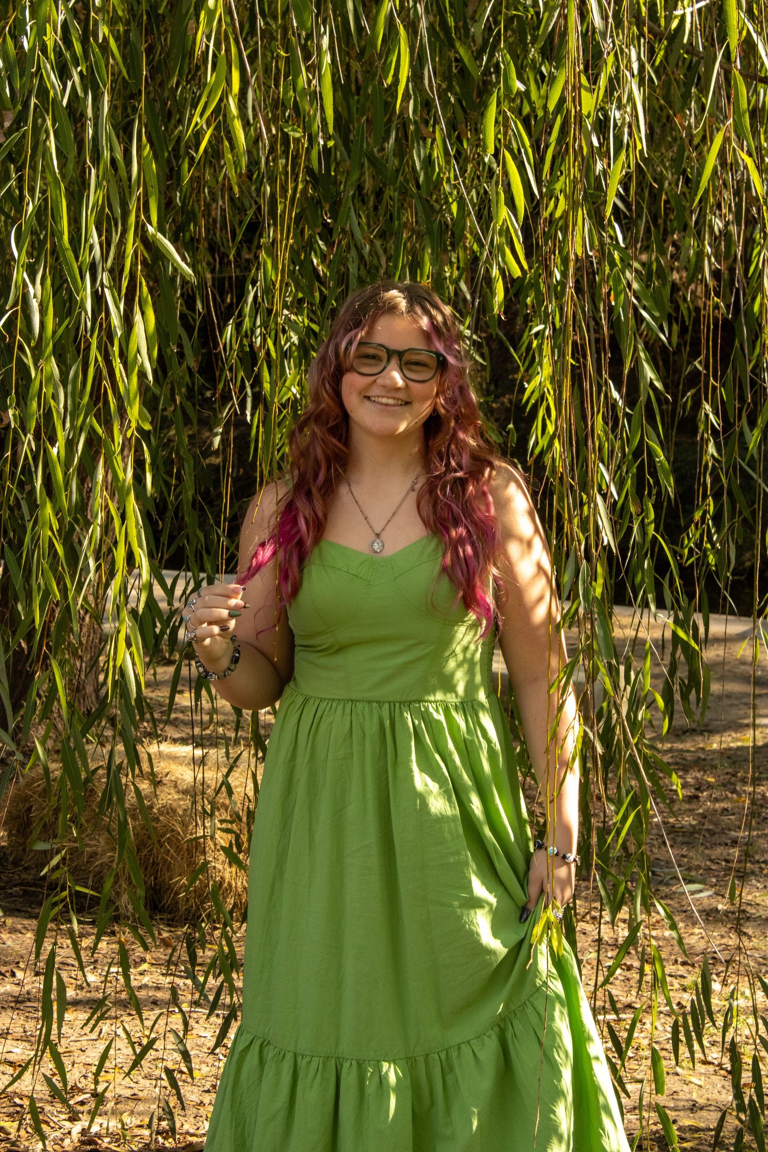 Someone wearing a bright lime green summer dress stands in dappled sunlight beneath drooping willow tree branches.