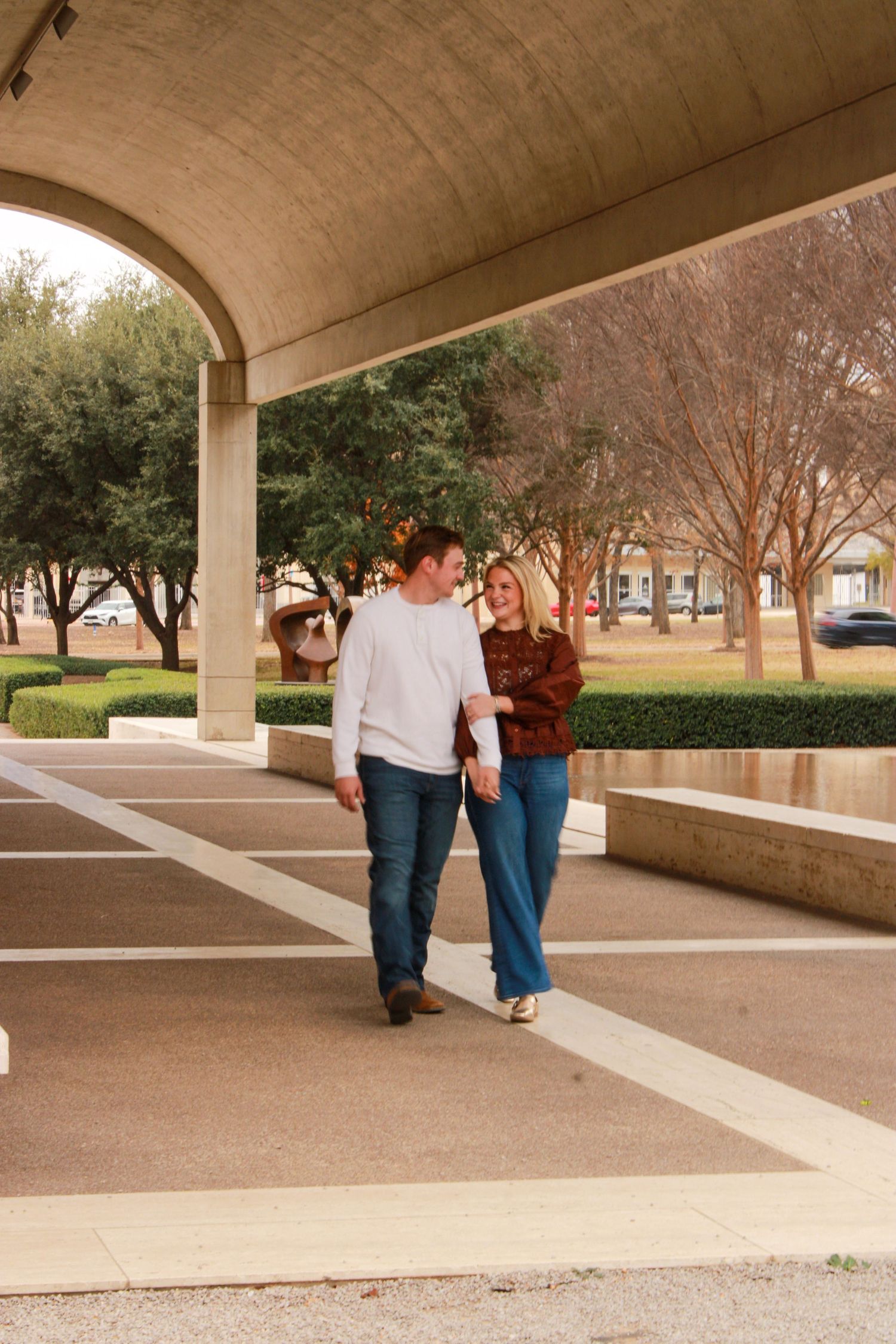 A couple walks together under a covered walkway on a college campus with trees in the background.