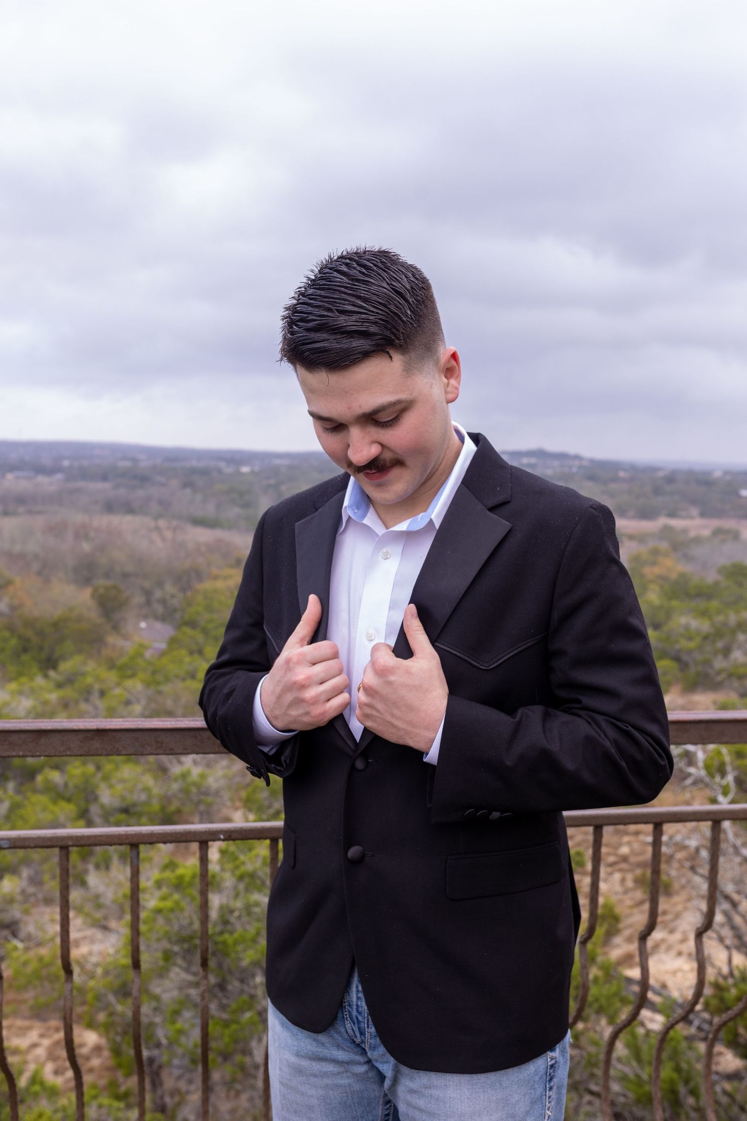 Young man with mustache adjusting black blazer over white shirt, standing on outdoor balcony with scenic landscape view.