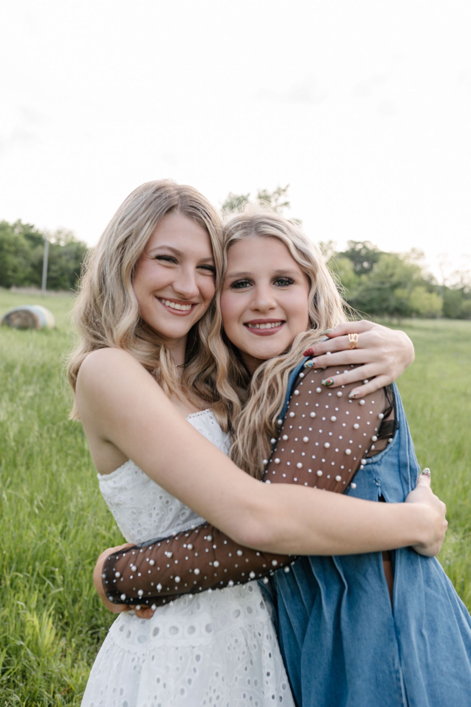 Two blonde women hugging and smiling in a green field, one wearing a white dress and the other a blue denim outfit.