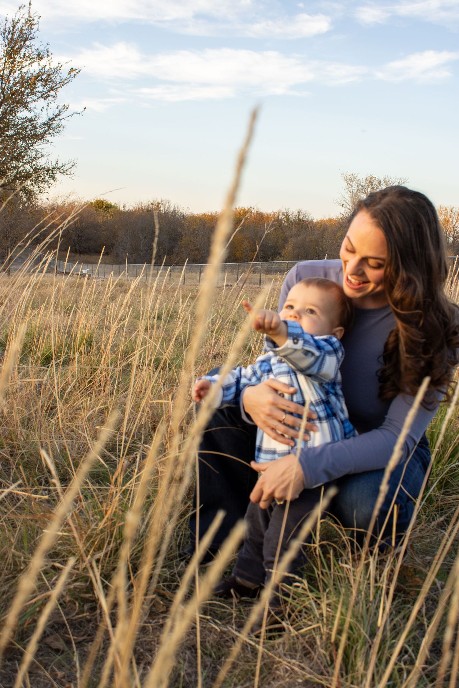 Mother and baby share joyful moments together while sitting in tall grass during golden hour.