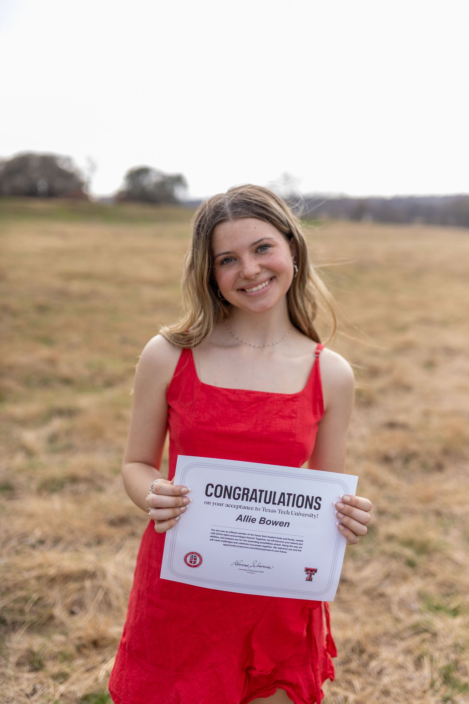 Someone in a red dress holds up a congratulations certificate while standing in an open field on a sunny day.