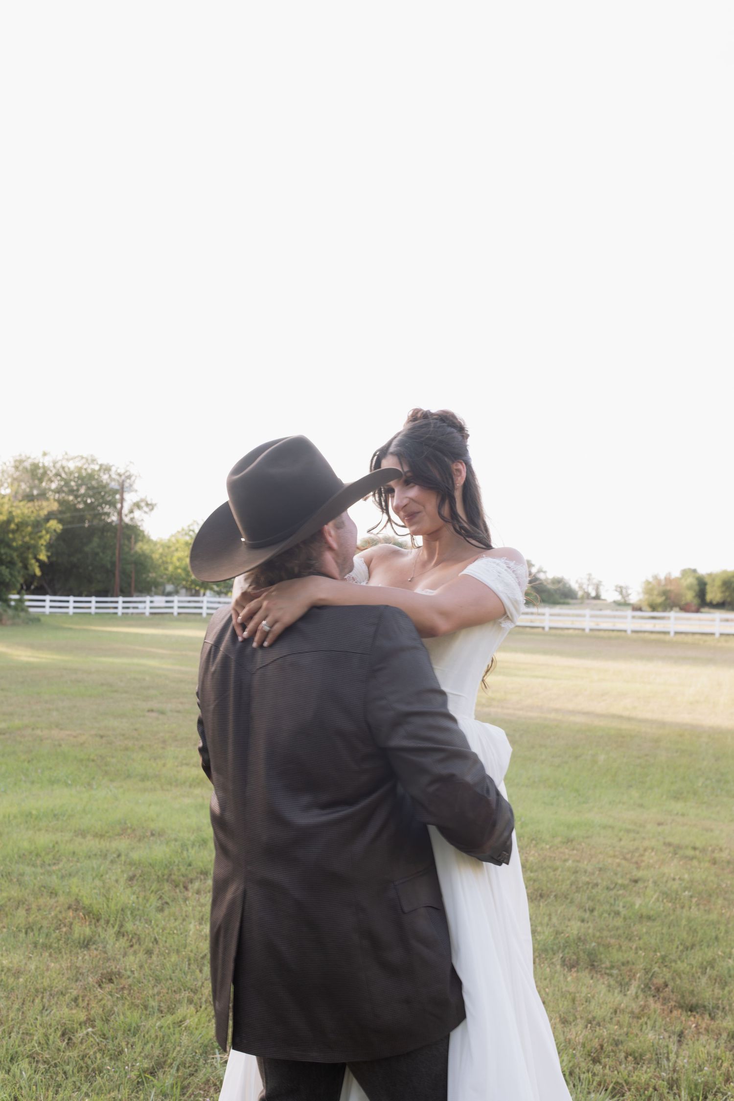 Bride in white gown smiling at groom in cowboy hat dancing in open field at sunset wedding.