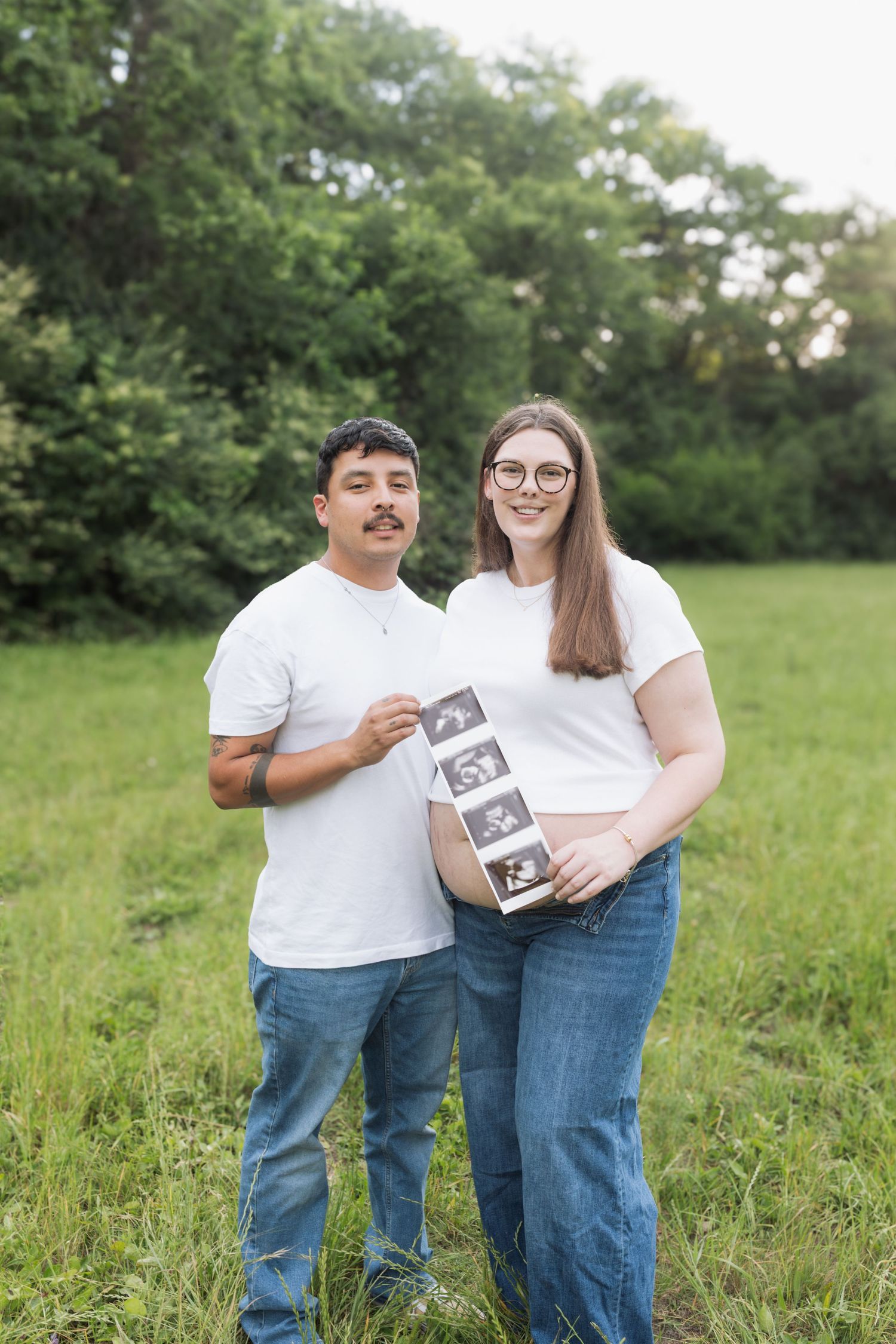 Expecting couple in white tees and jeans holding ultrasound photos in a green field, announcing pregnancy outdoors.