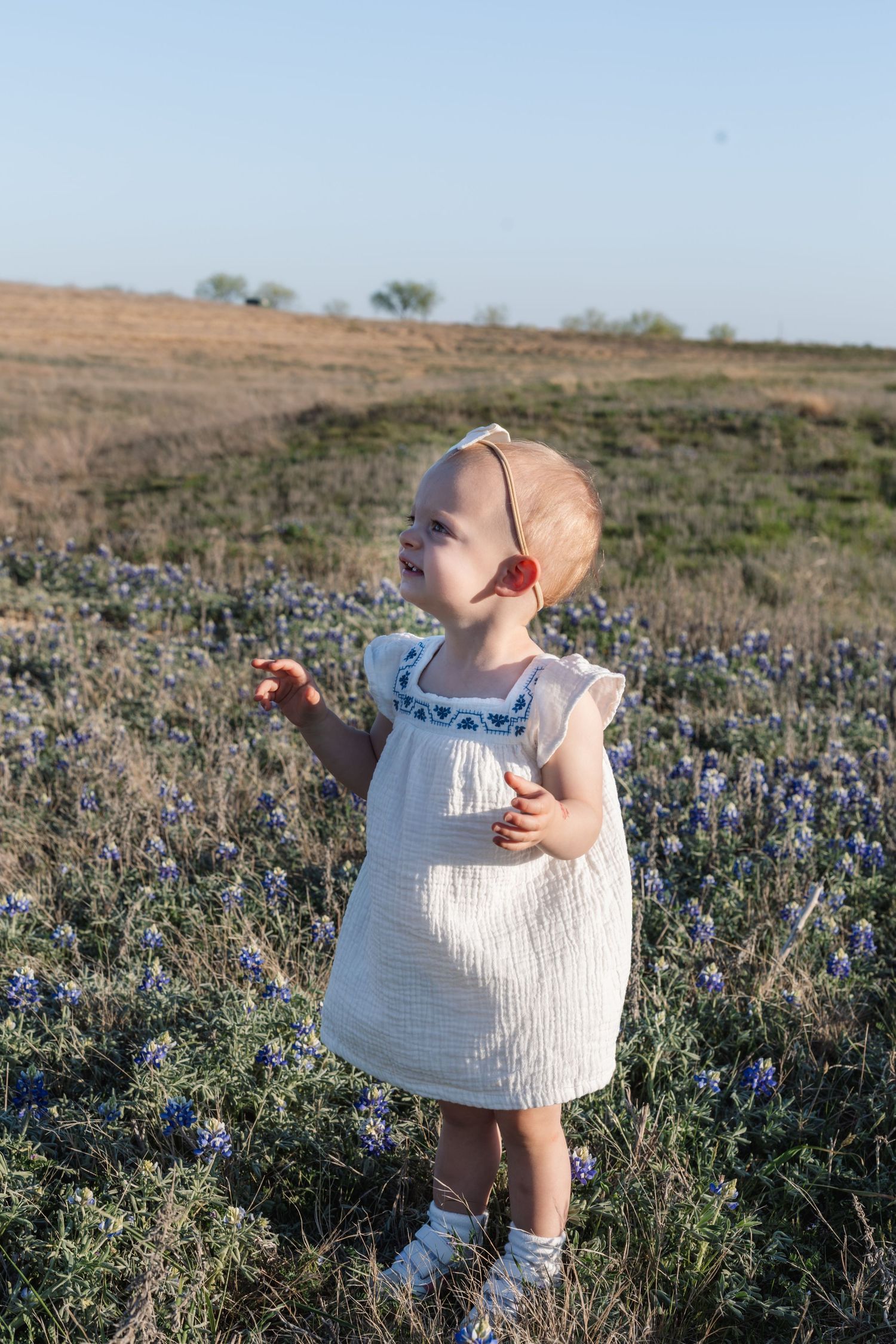 Small child in white dress stands in a wildflower field at sunset holding something while looking up at the sky.