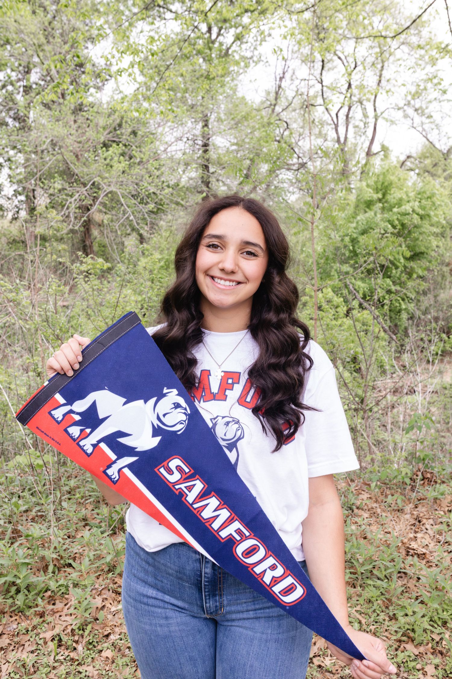 Student in casual attire holds school spirit banners while posing outdoors among trees and foliage.