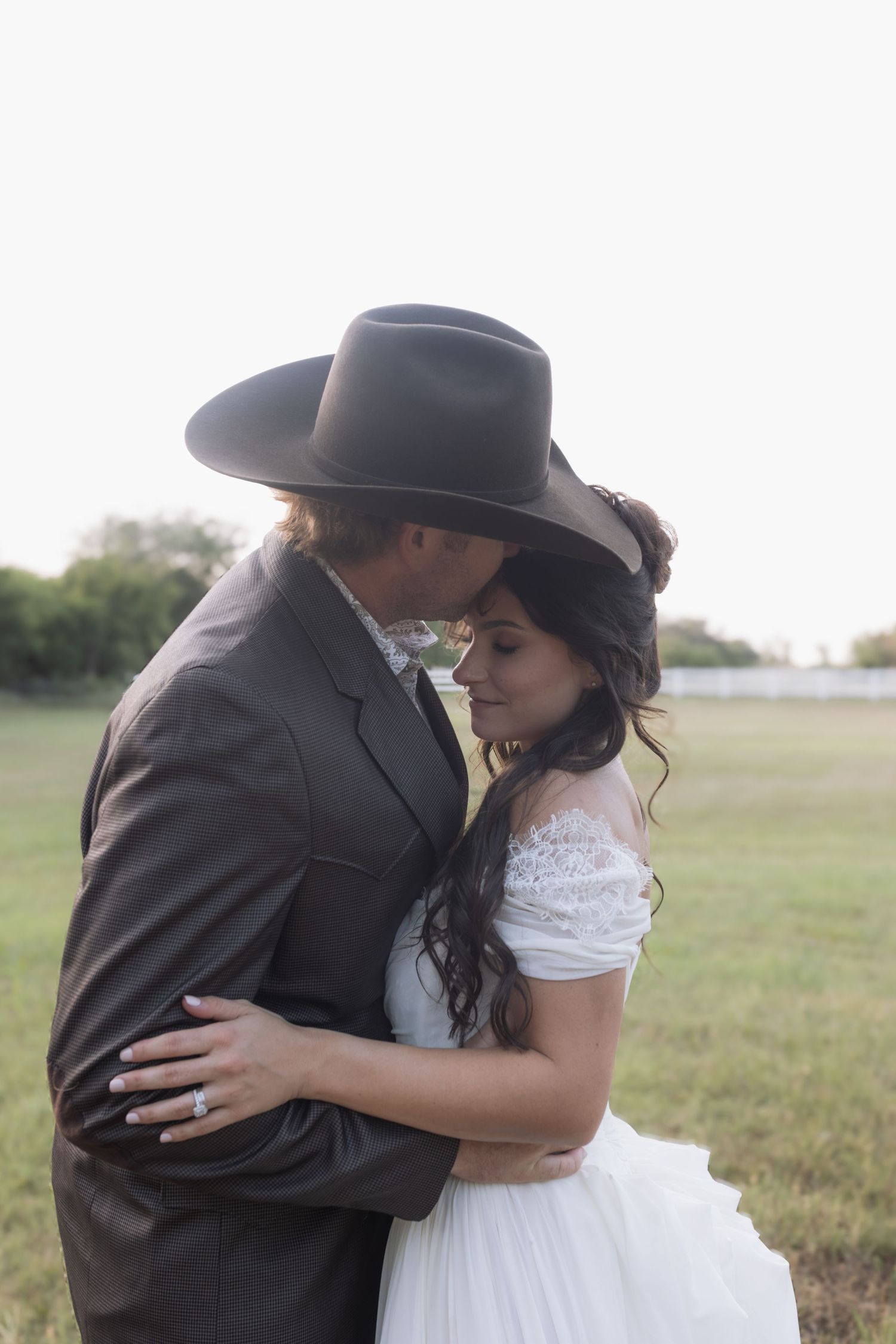 A bride and groom share a romantic moment outdoors, with the groom wearing a cowboy hat and the bride in a white lace dress.