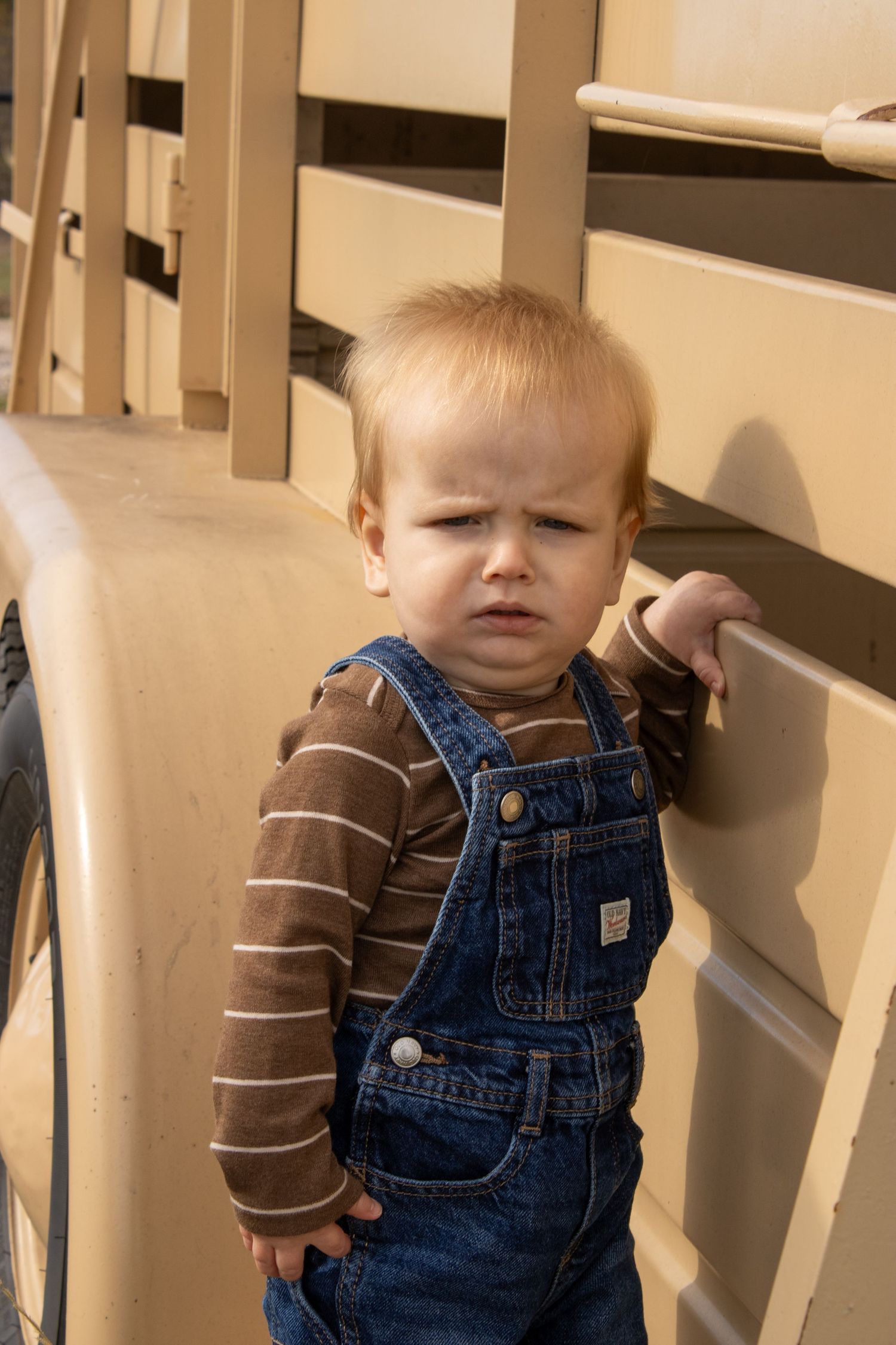 Small child wearing denim overalls and brown striped shirt stands against beige wall.