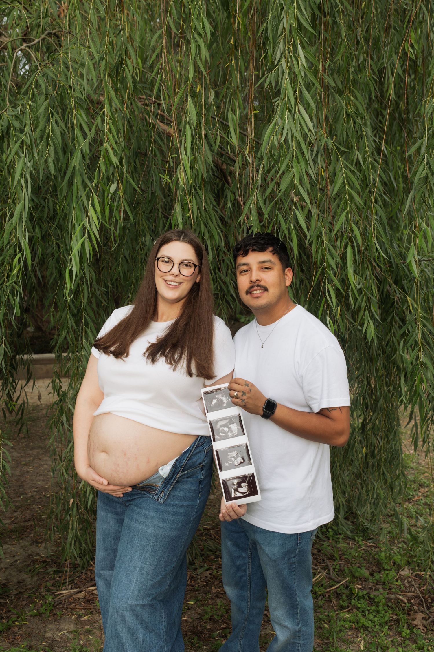 Happy expectant couple holding ultrasound photos under weeping willow tree, wearing matching white shirts and jeans.