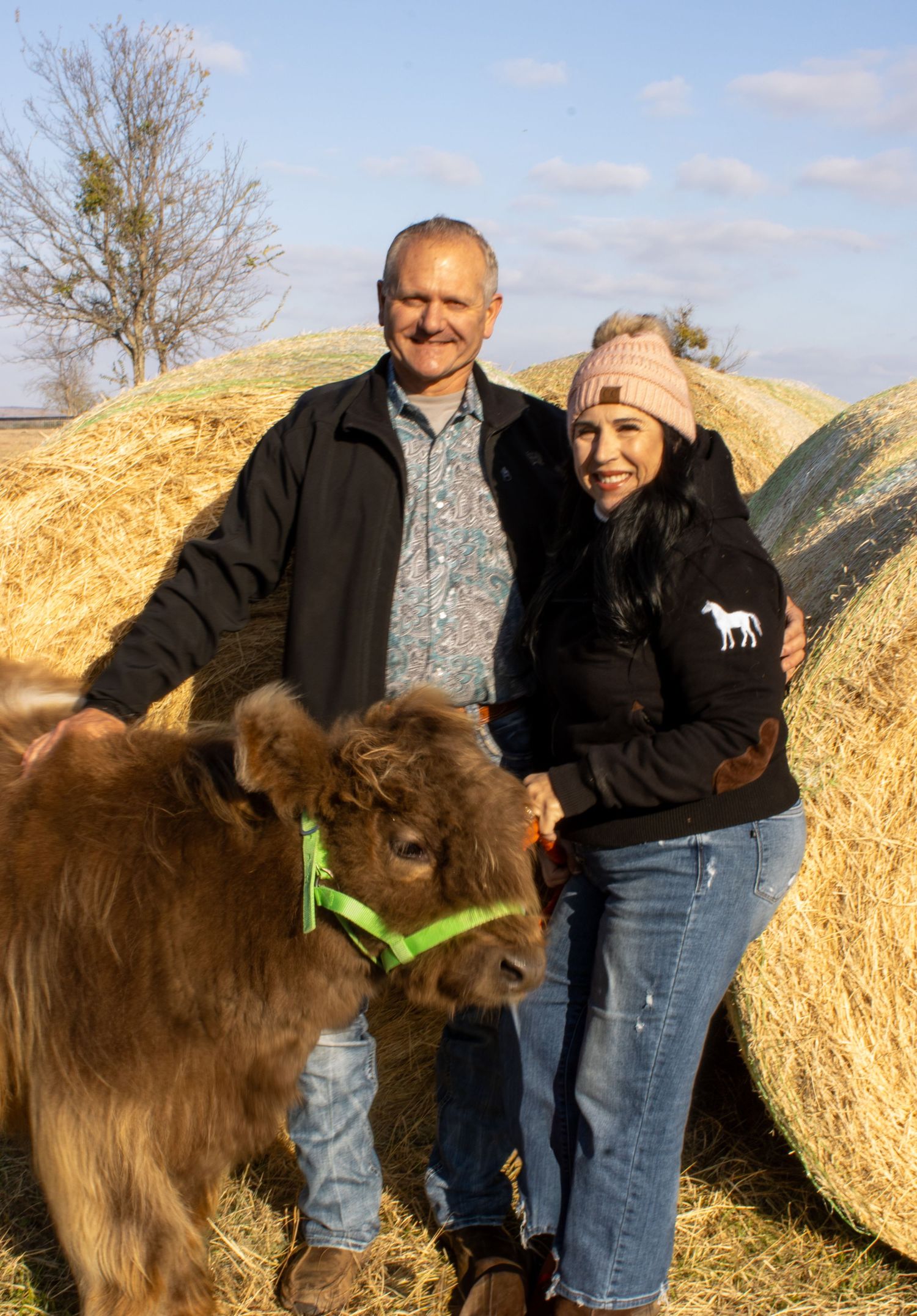 Two people stand with a large brown Newfoundland dog against hay bales in a sunny outdoor setting.