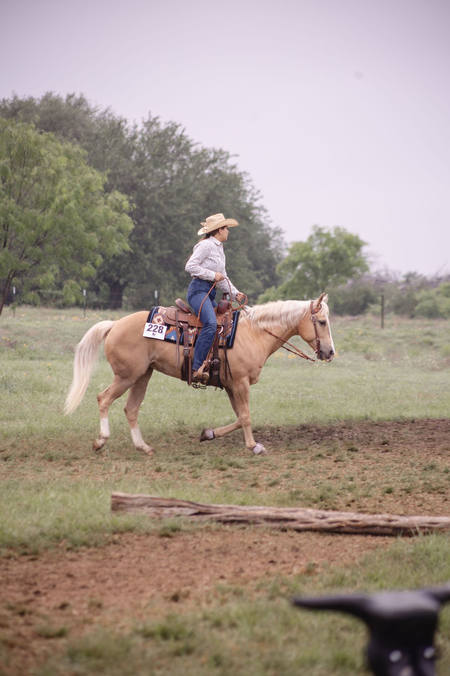A cowgirl in a hat rides a palomino horse in a grassy field during an equestrian competition.