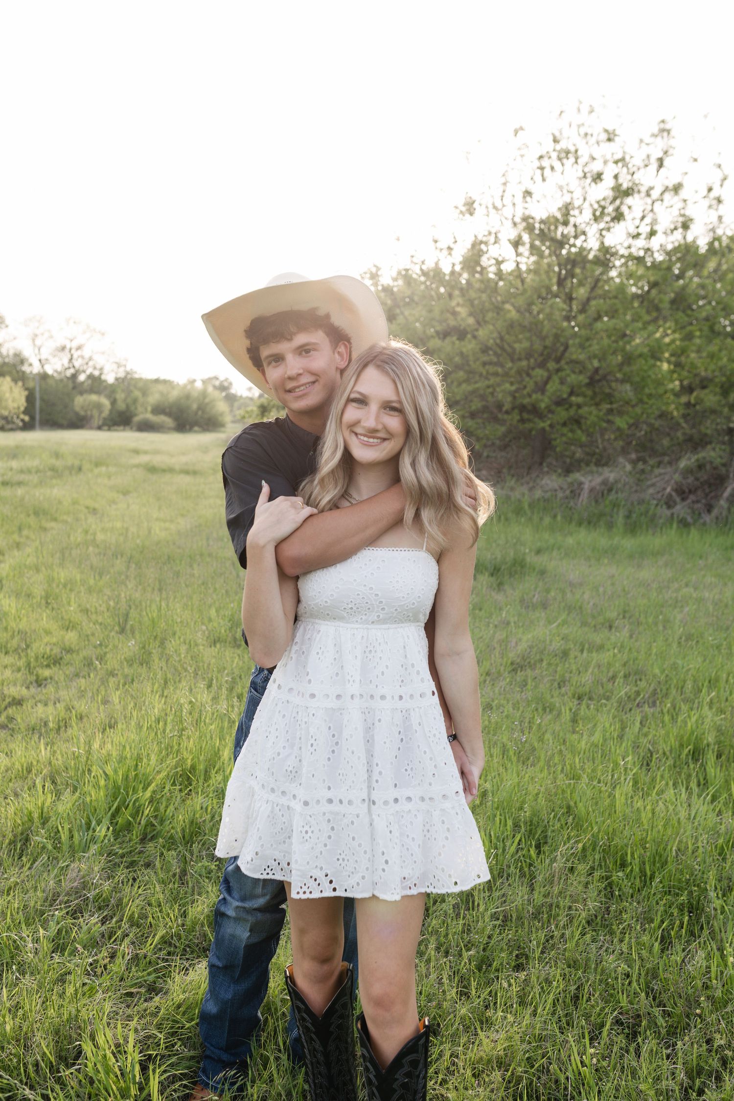 Young couple posing in a green field, woman in white dress and cowboy boots, man in cowboy hat embracing her.