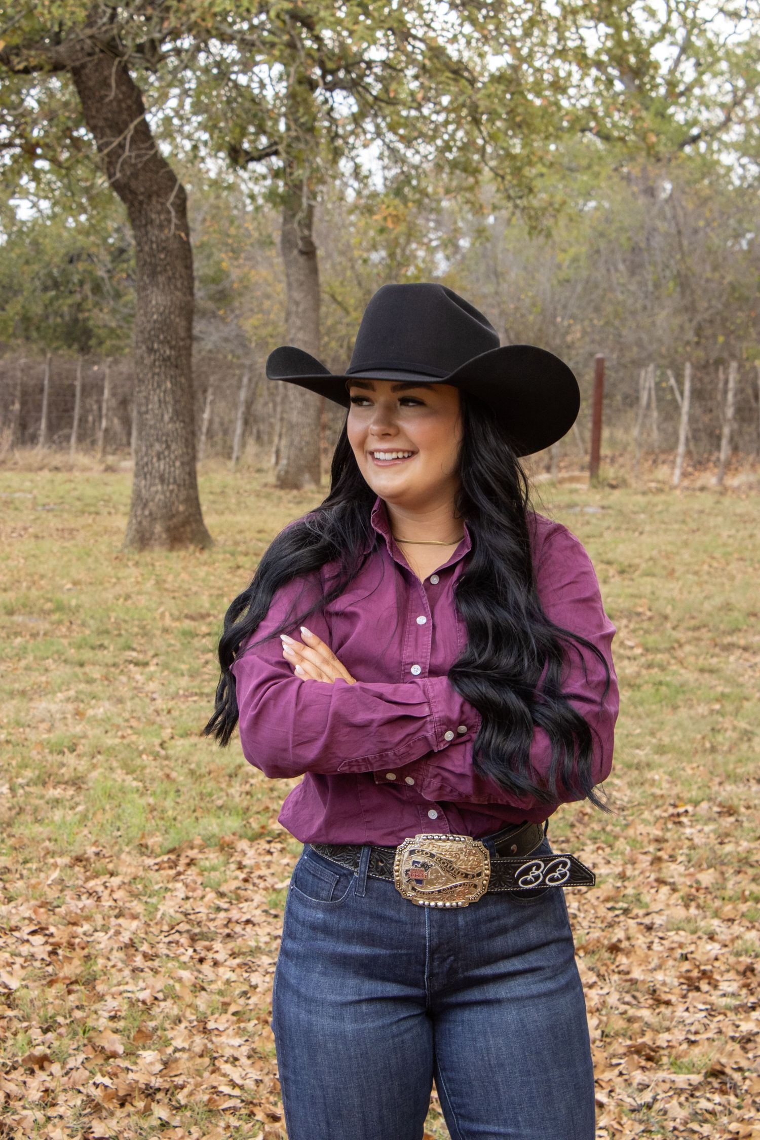 A person in a purple western shirt and wide-brimmed black hat poses outdoors among autumn trees and fallen leaves.