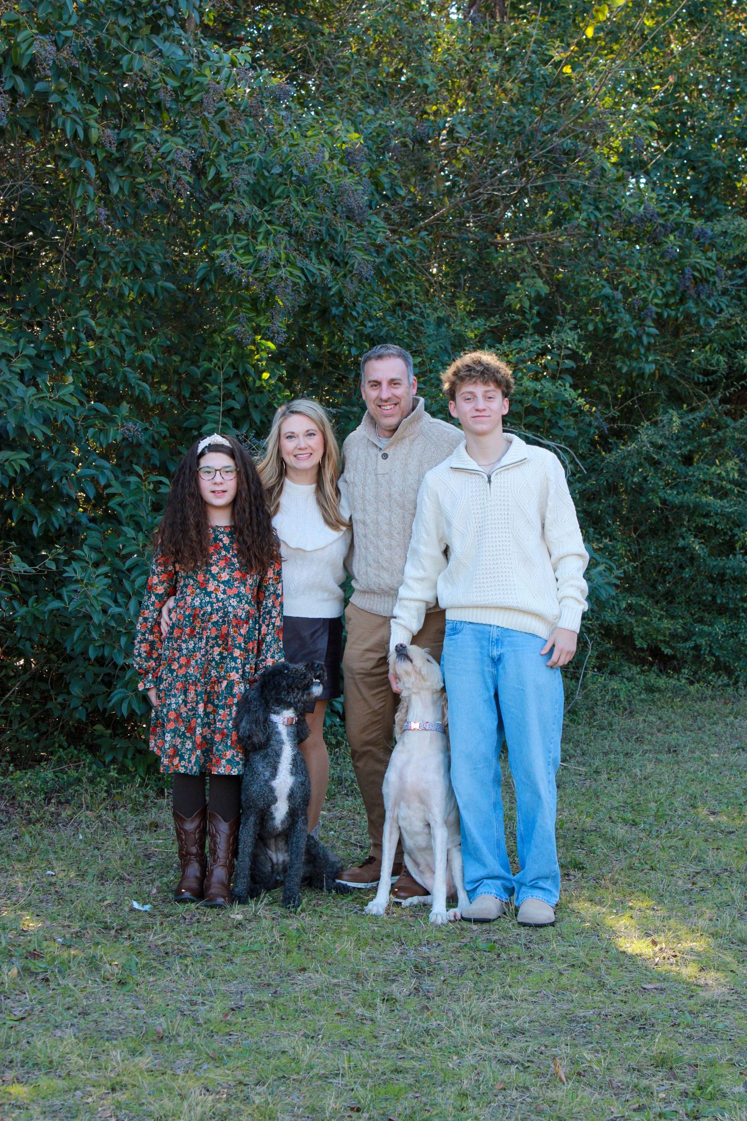 A group poses outdoors with two dogs against a green foliage backdrop on a sunny day.