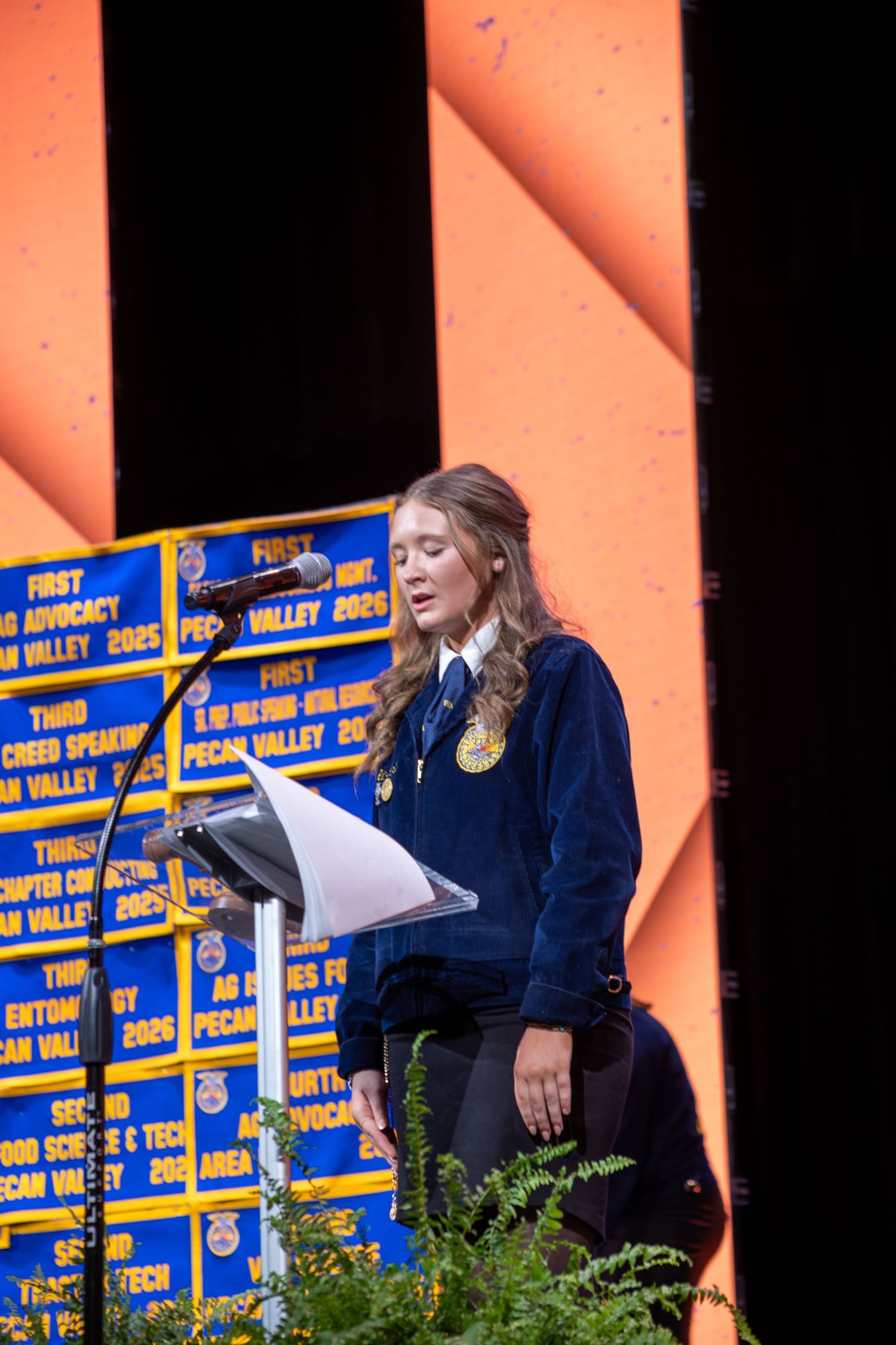 Young FFA member in blue jacket speaks at podium during awards ceremony with blue banners displayed behind her.