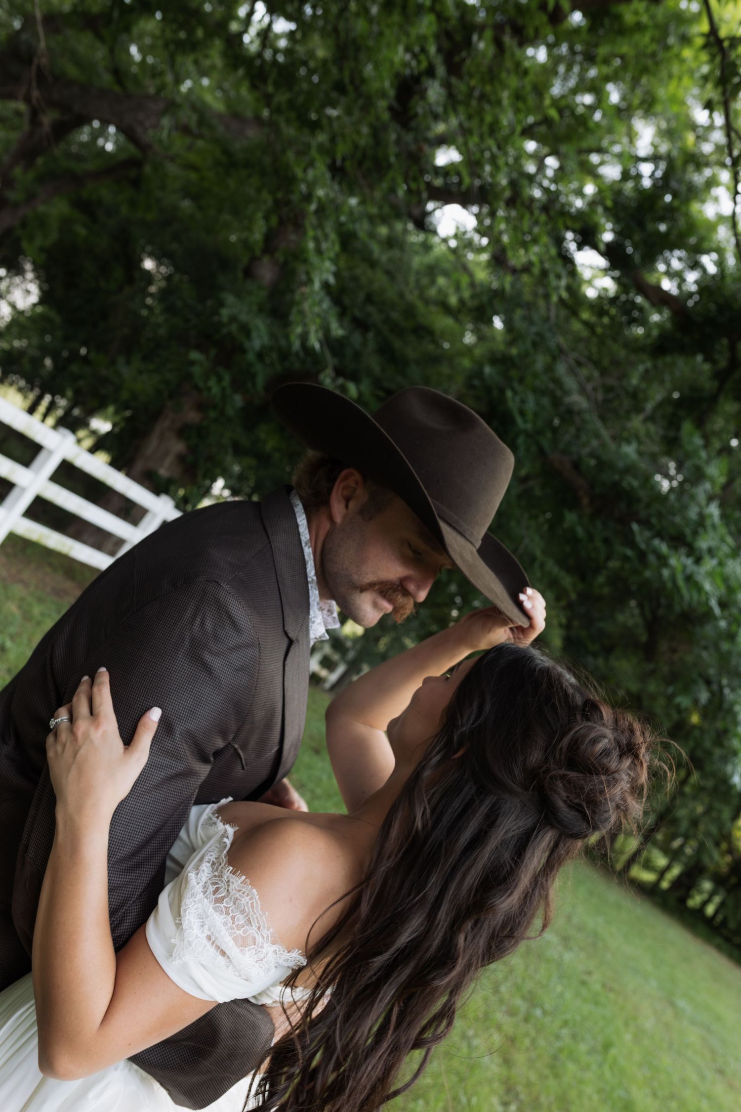 Bride and groom sharing a romantic kiss outdoors, groom in cowboy hat and dark suit near white fence.